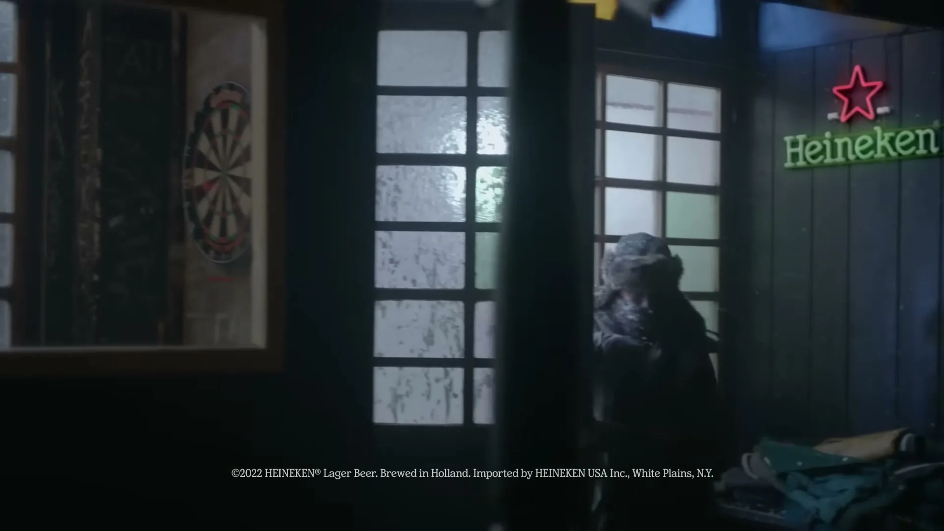 A person in a winter coat enters a dark pub with a dartboard and a Heineken neon sign, as snow falls outside.