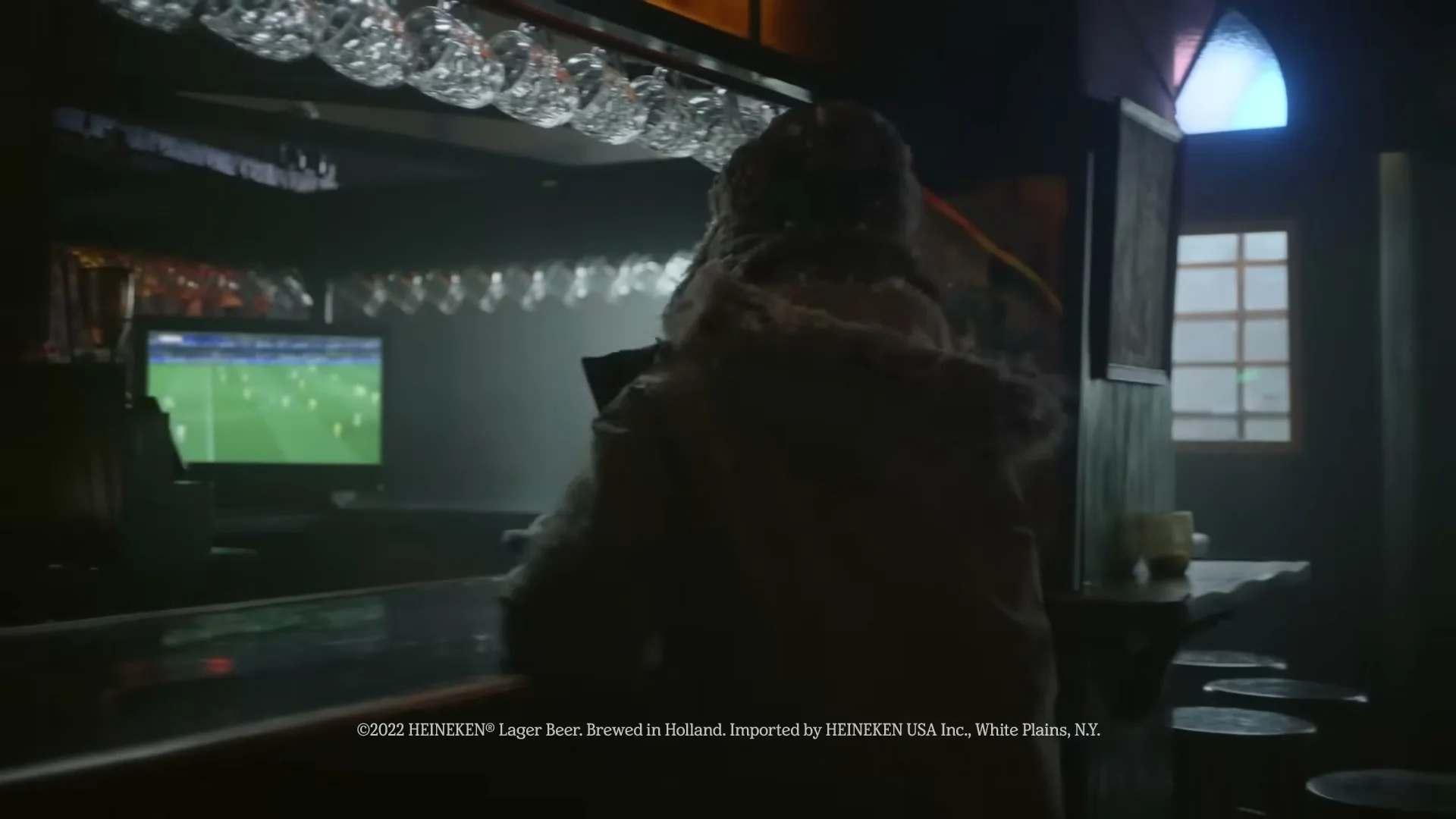 A person in a furry hat and winter coat sits at a bar, watching a soccer game on a TV in a dimly lit pub.
