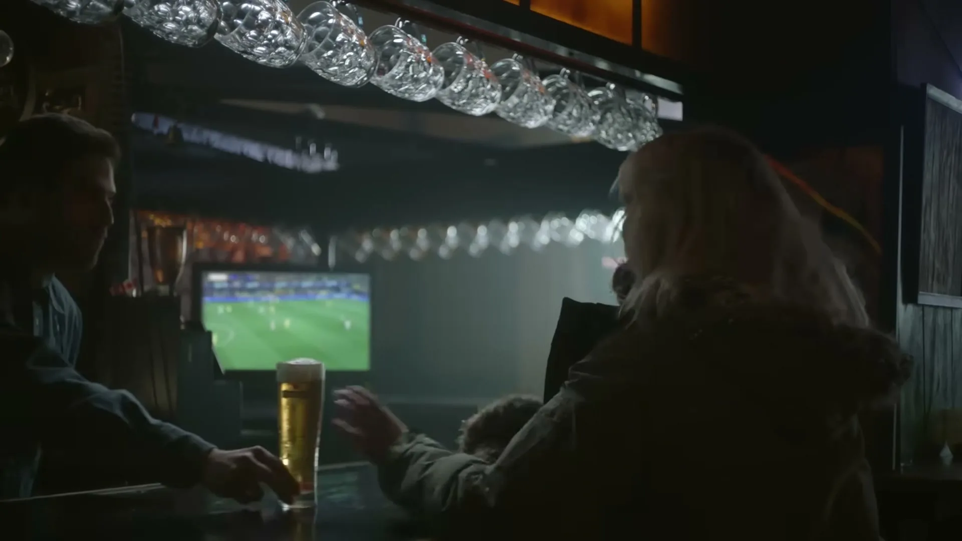 A blonde woman in a winter coat sits at a bar, receiving a glass of beer from a male bartender, with a soccer game playing on a TV in the background.