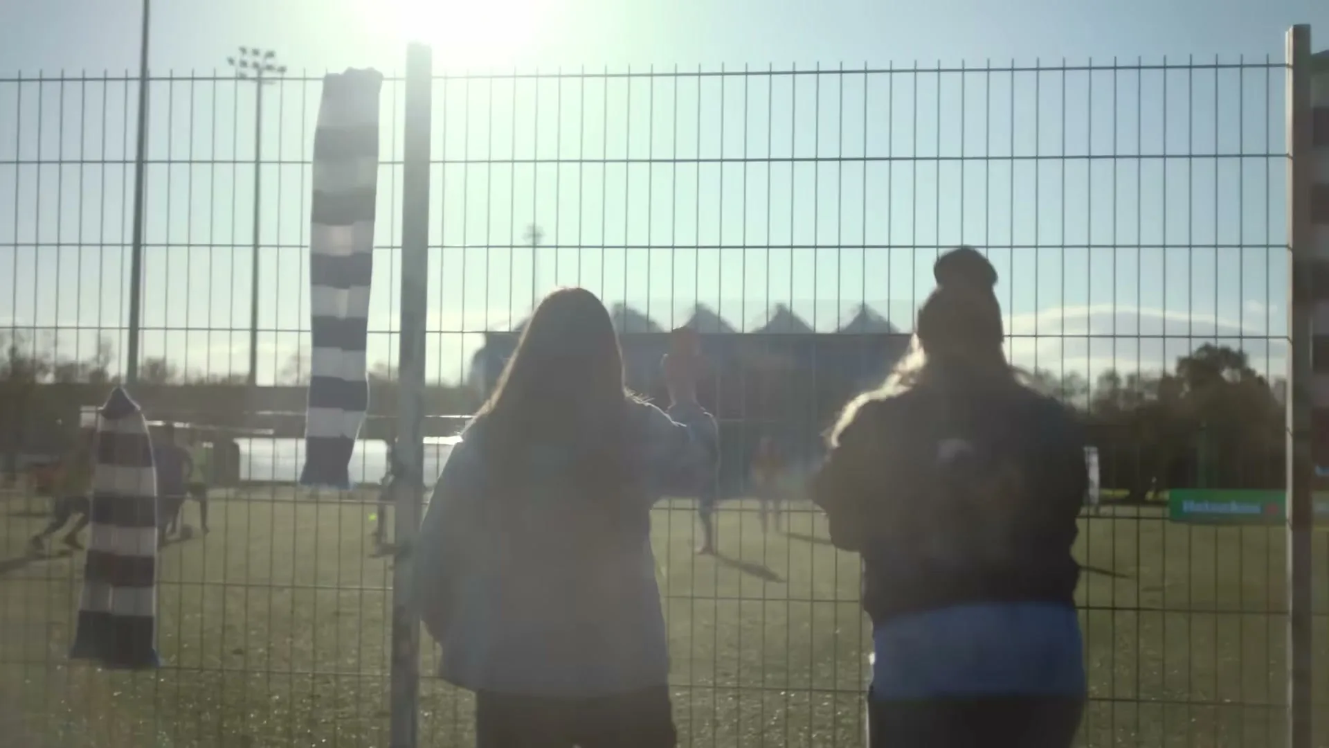 Two women with long hair watch a soccer game from behind a fence on a sunny day, with a stadium in the background.
