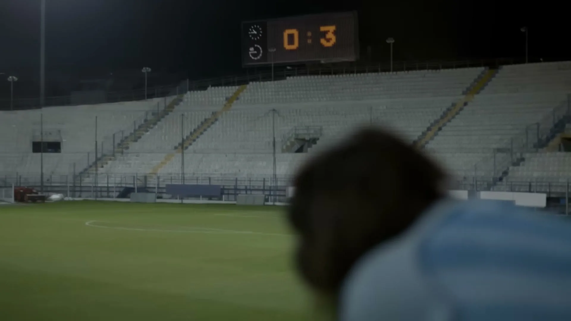 A blurry male soccer player in a light blue jersey watches the scoreboard showing "0:3" in an empty stadium at night.