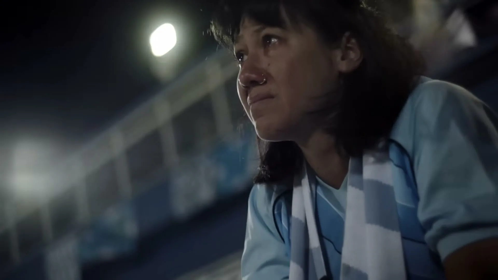 A female soccer fan with a nose ring, wearing a light blue jersey and scarf, looks on with tears in her eyes at a night stadium.