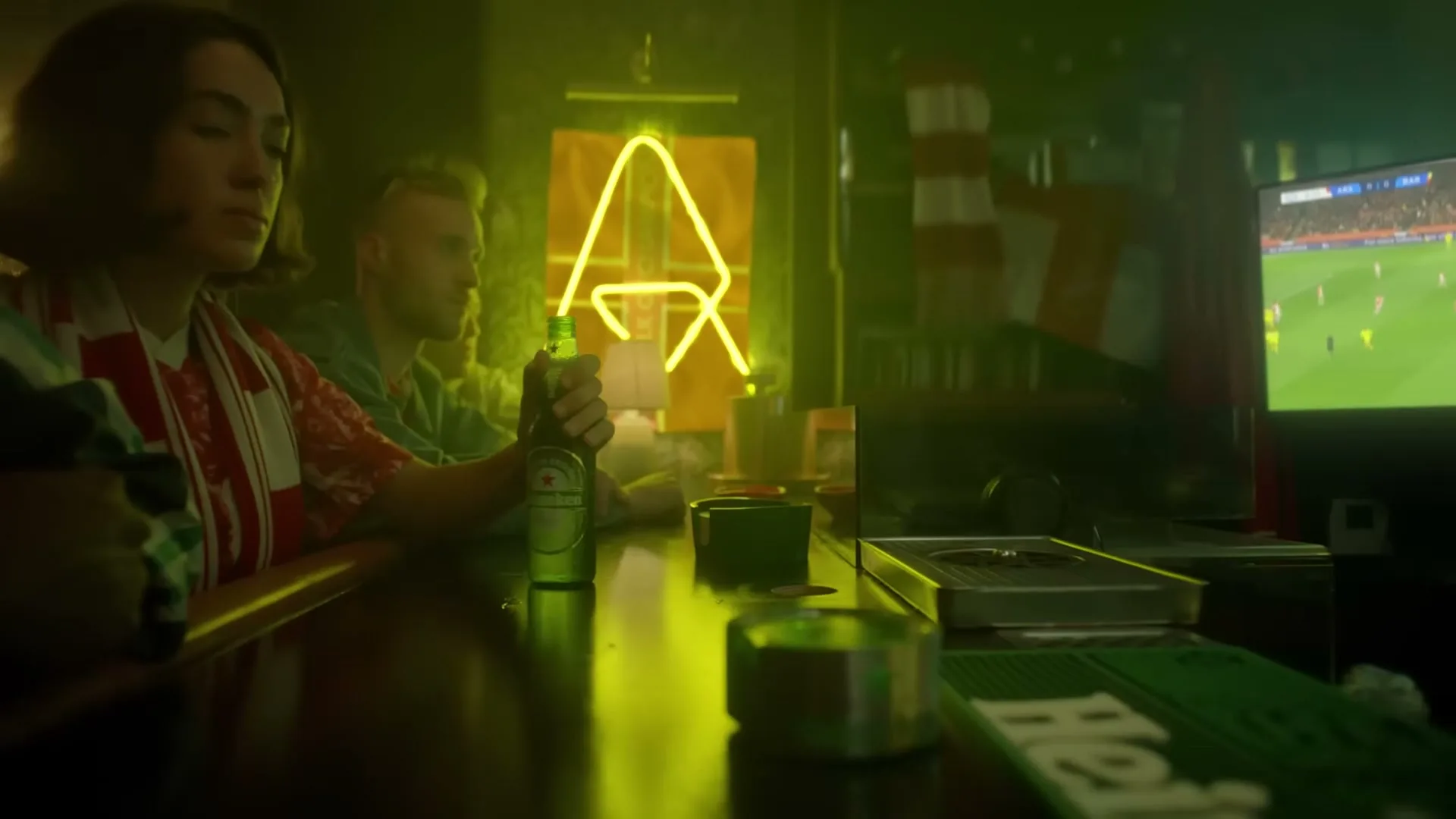 A diverse group of people watch a soccer game on TV in a dimly lit bar, as a hand slides a Heineken bottle across the counter.