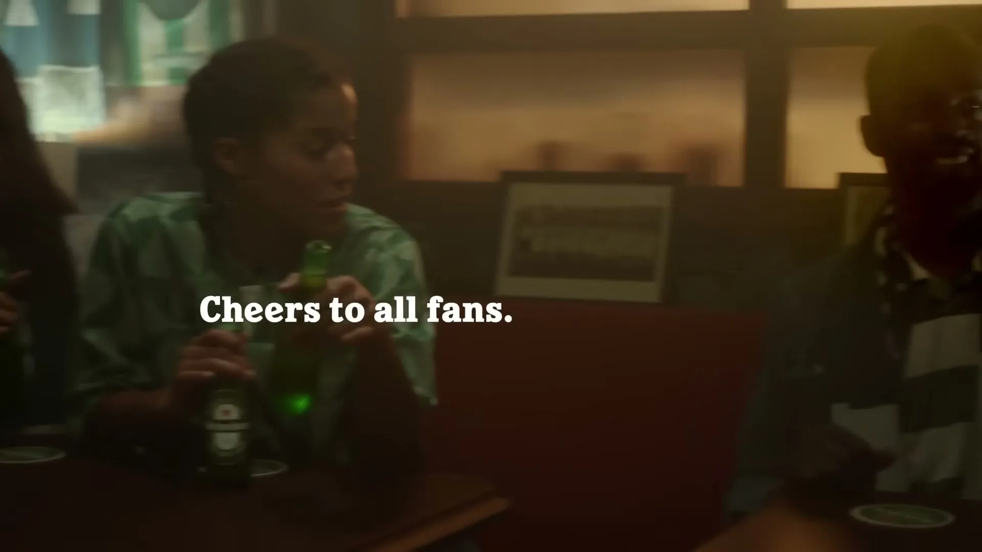 Three women in green and white shirts are sitting at a table in a dimly lit bar, talking and drinking Heineken beer.