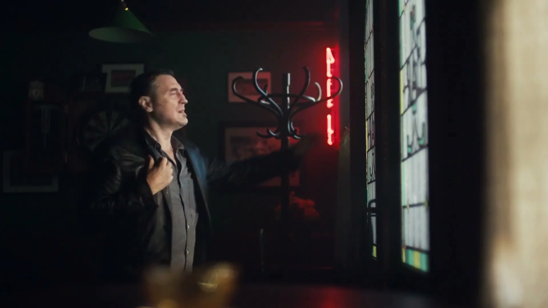 A man in his 40s, wearing a black leather jacket, stands in a dimly lit pub, looking out a stained-glass window.