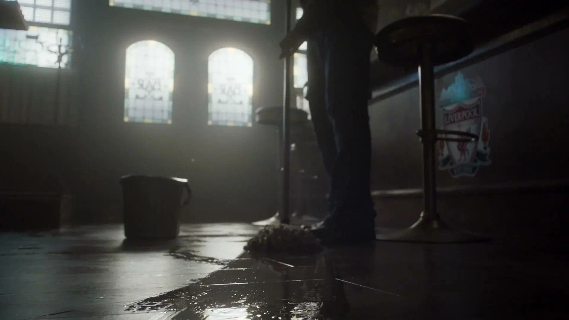 A person mops a wet floor in a dimly lit bar, with light streaming through stained glass windows in the background.