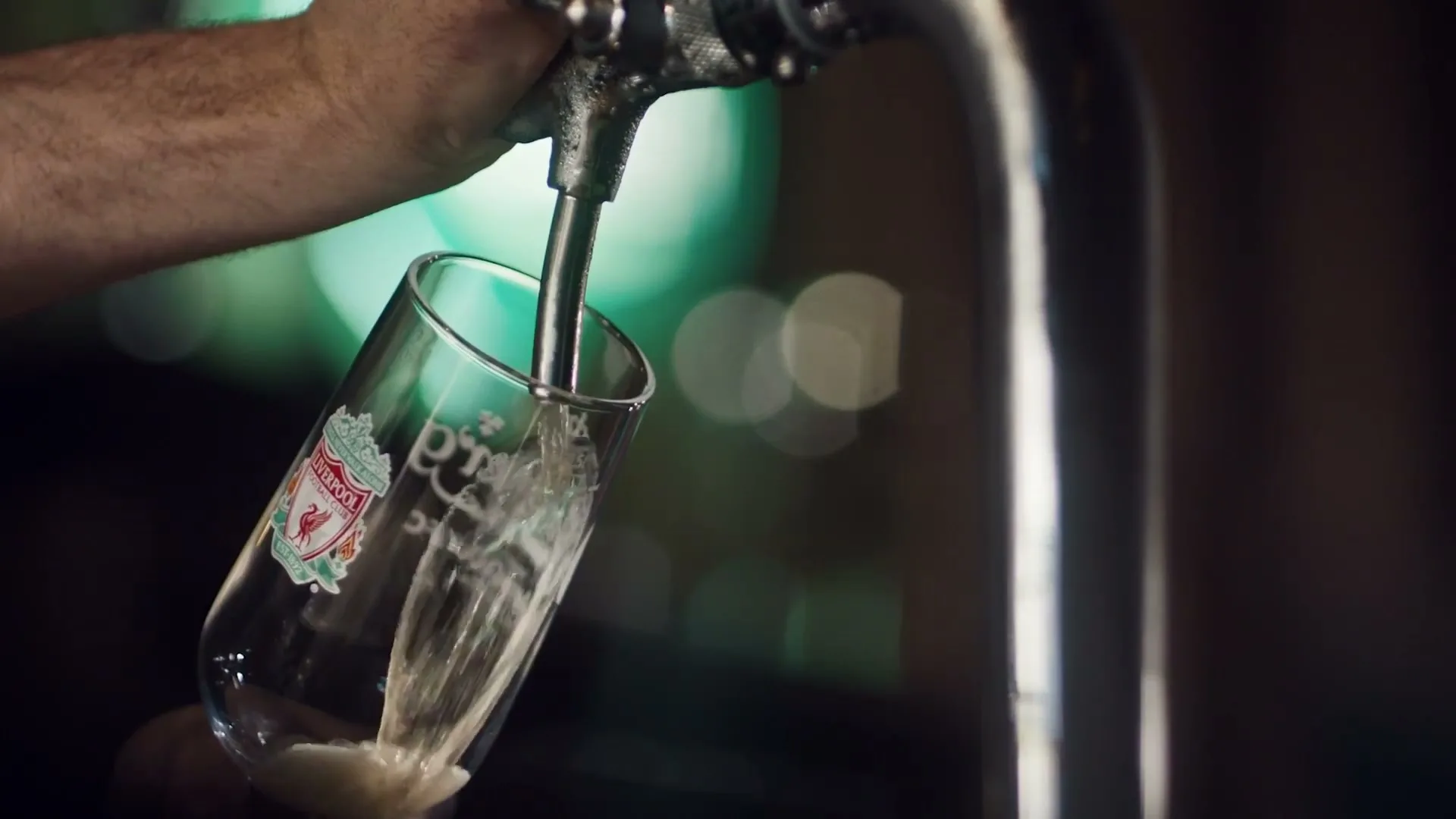 A bartender's arm holds a Liverpool FC branded glass under a beer tap, filling it with beer in a dimly lit pub.