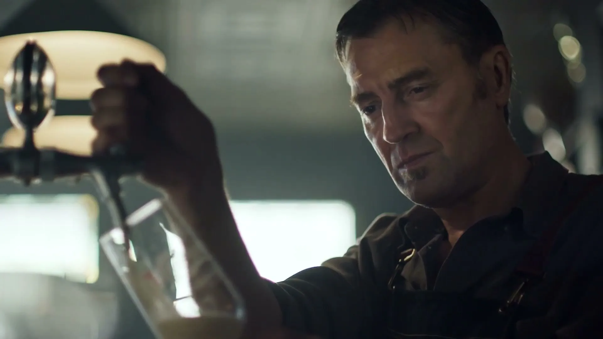 A male bartender in his 40s, wearing a dark shirt and suspenders, intently pours a beer from a tap into a glass in a dimly lit bar.