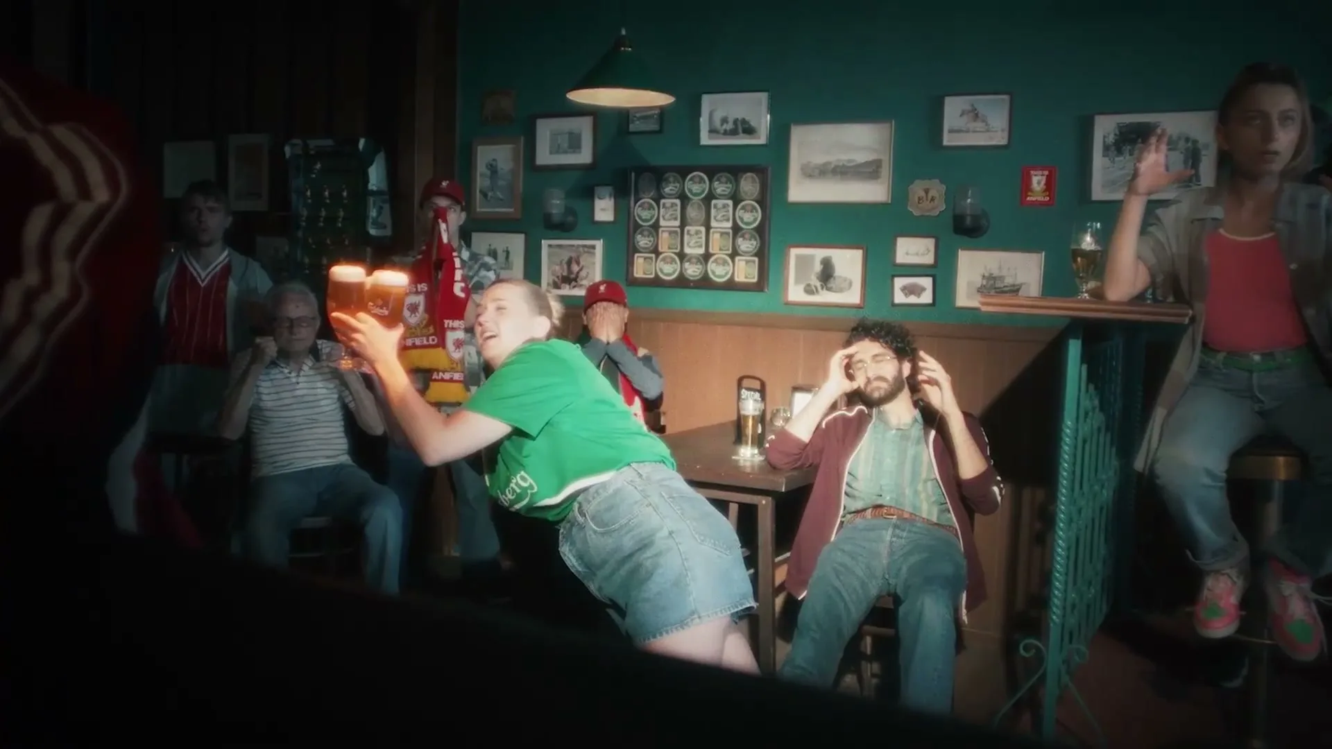A group of football fans in a pub react to a game, with one woman holding up two pints of beer in excitement.