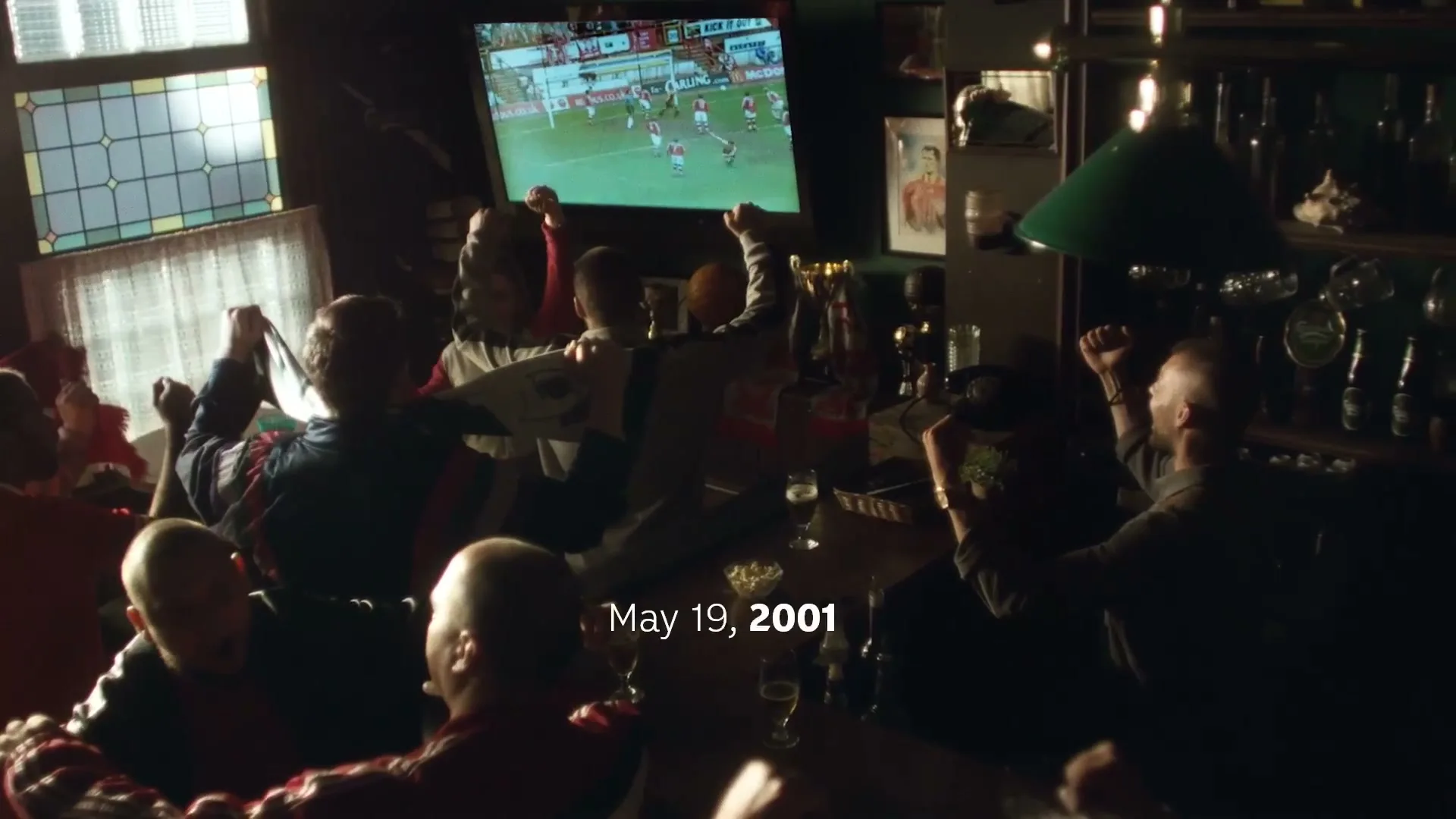 A group of male football fans in a dimly lit pub cheer as they watch a match on a TV screen, with some raising their arms in excitement.