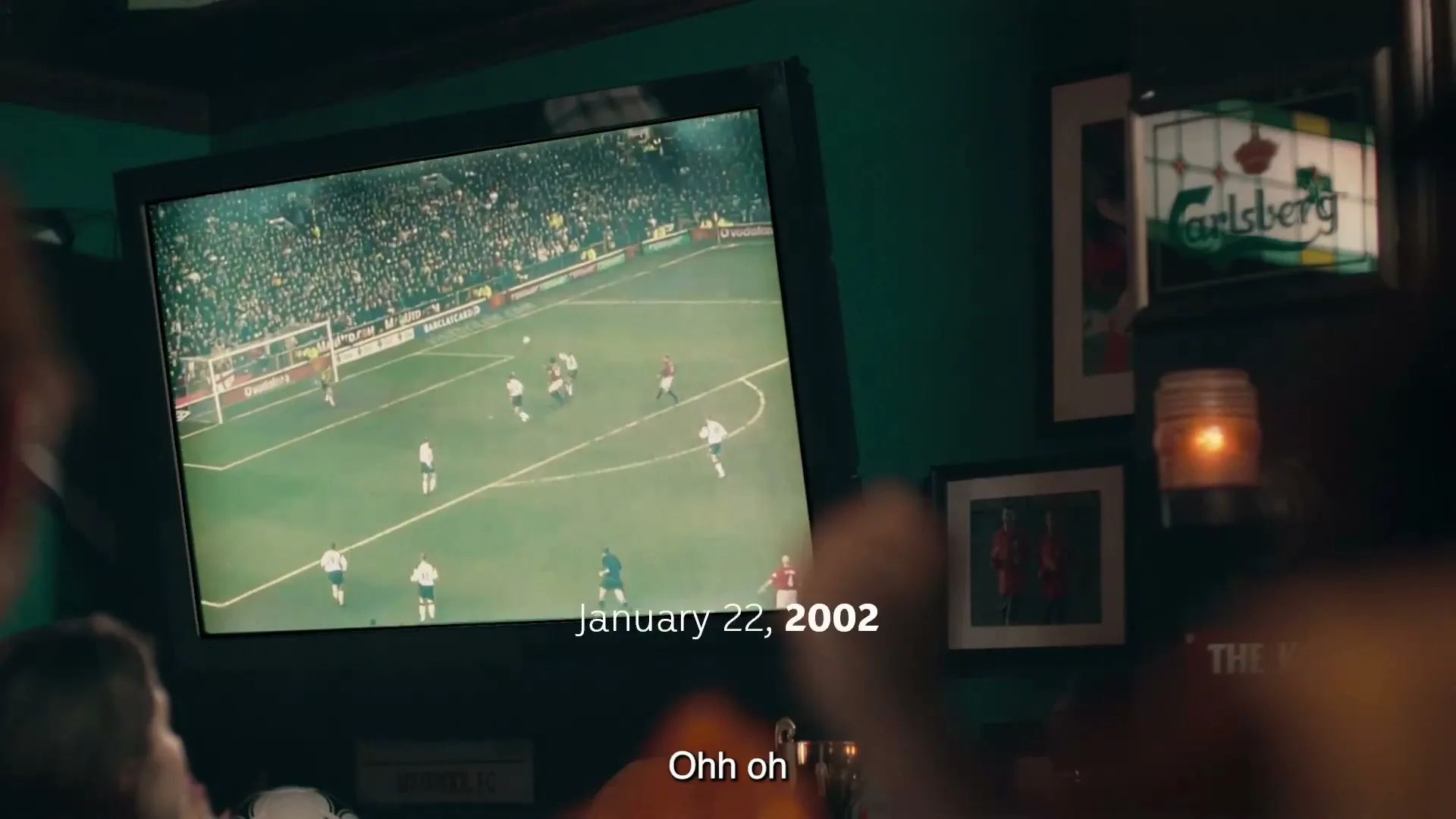 A group of people watch a soccer game on a TV screen in a dimly lit pub, with a Carlsberg sign visible on the wall.
