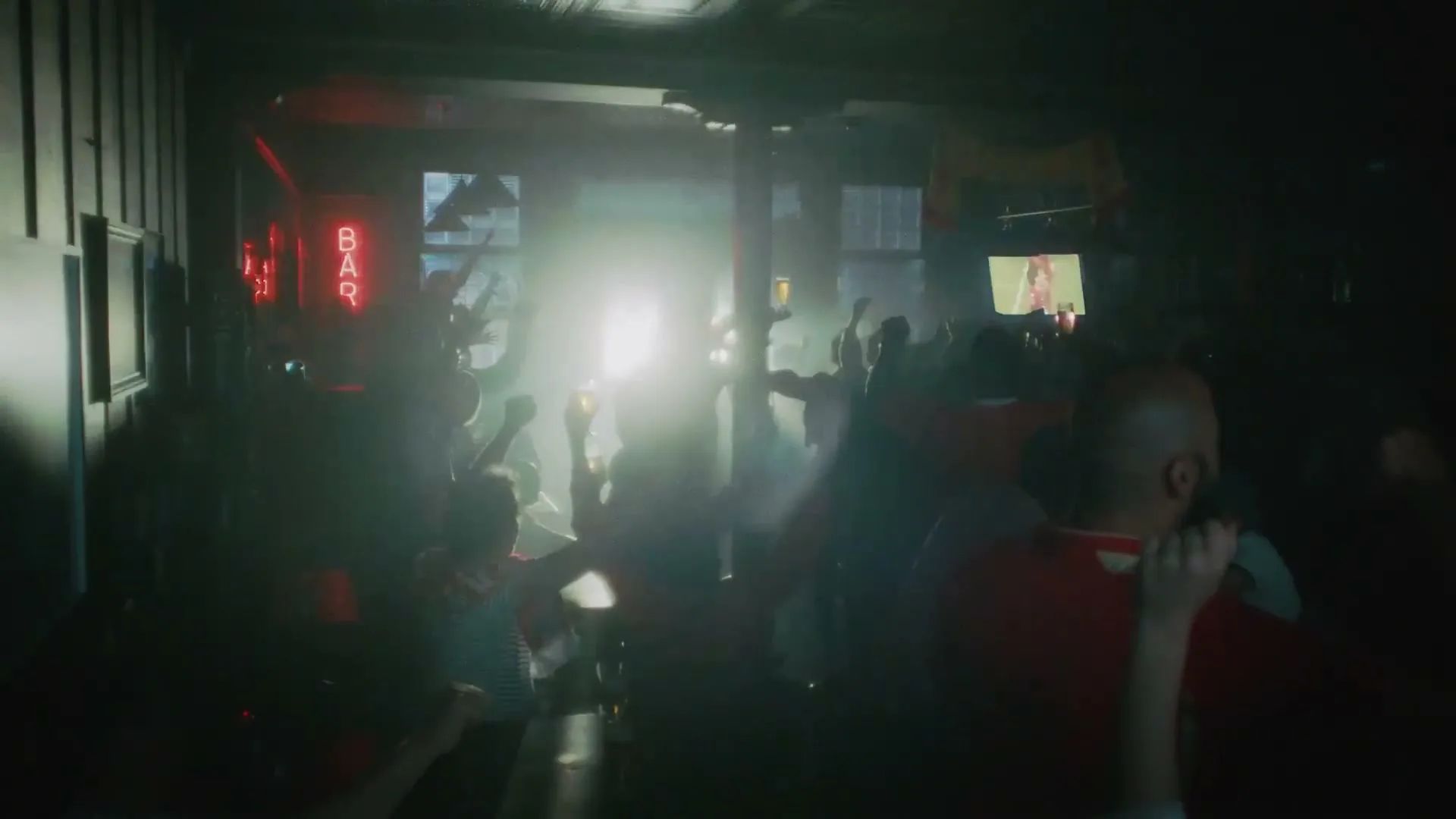 A diverse crowd of football fans in a dimly lit bar cheers and raises their arms, celebrating a goal on a TV screen.