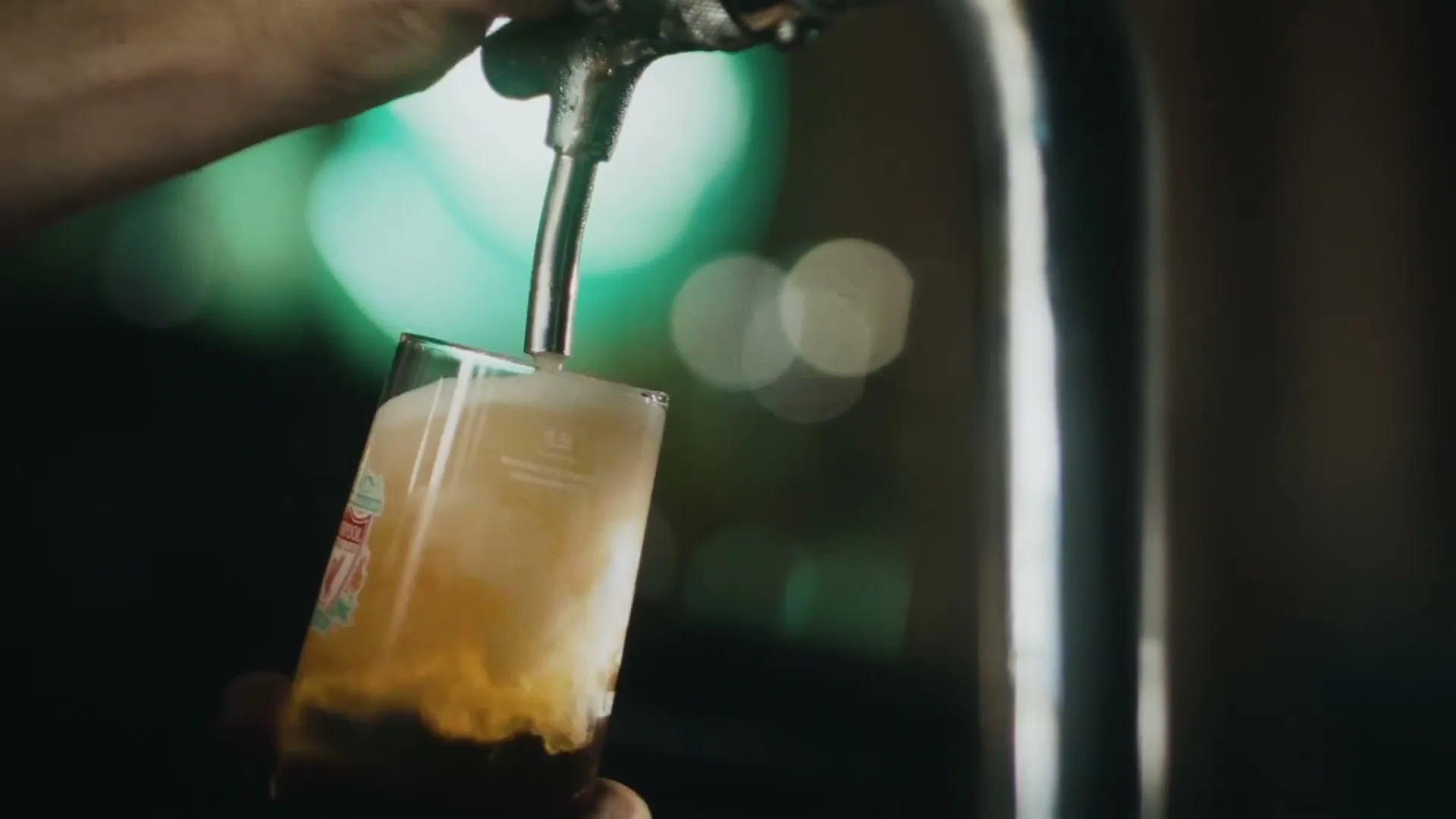 A bartender's hand holds a glass with a Liverpool FC logo as beer is poured from a tap, filling the glass with foam in a dimly lit pub.