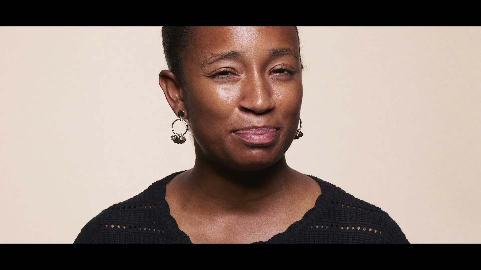 A Black woman with short hair and a black top looks directly at the camera, making a slightly uncomfortable face against a plain background.