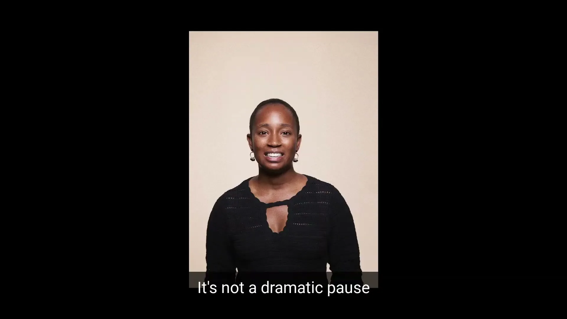 A Black woman in her 30s, wearing a black top, smiles and speaks directly to the camera against a plain beige background.