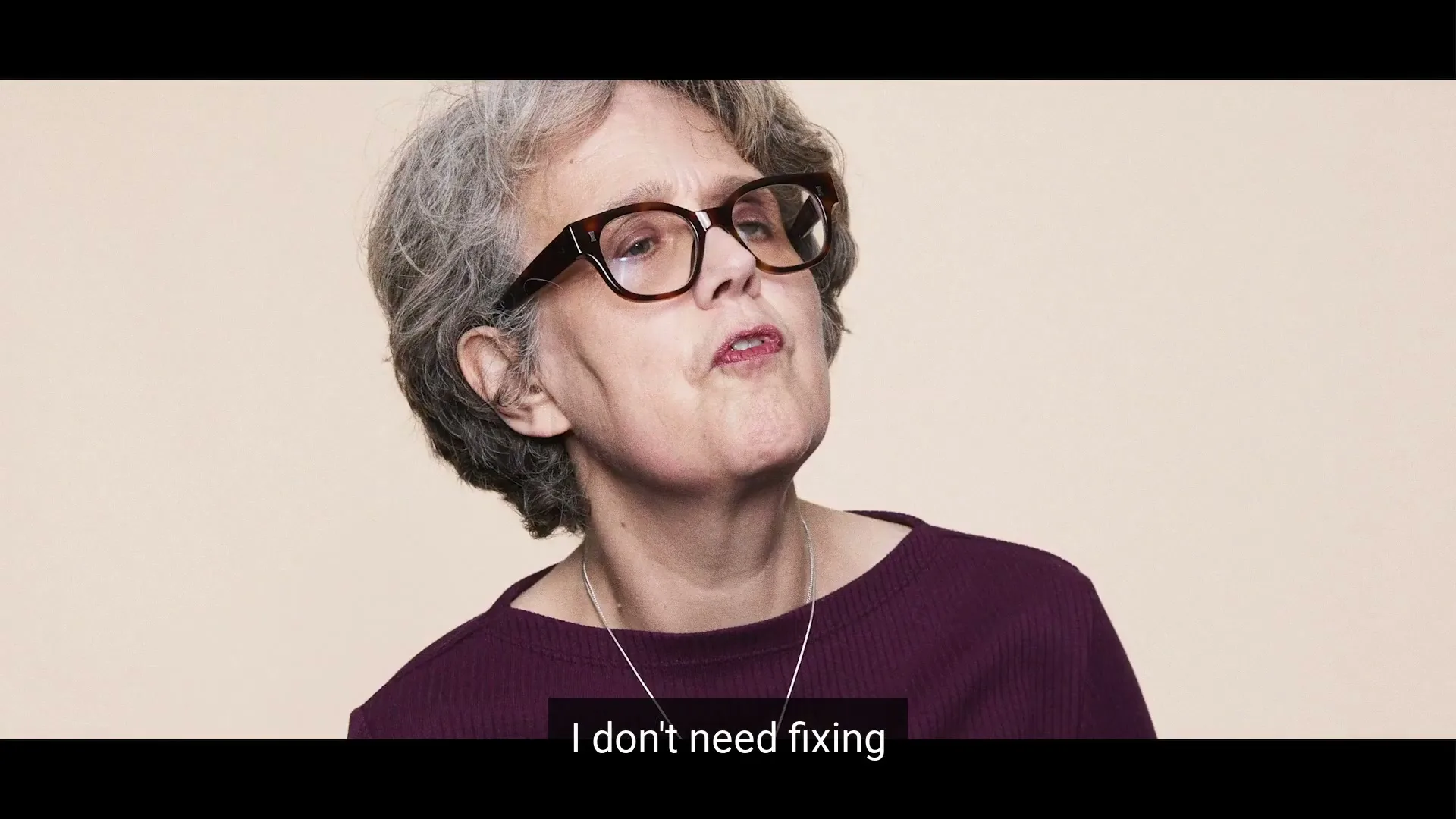 A woman with grey hair and glasses looks up and to the right, speaking with a determined expression against a plain background.