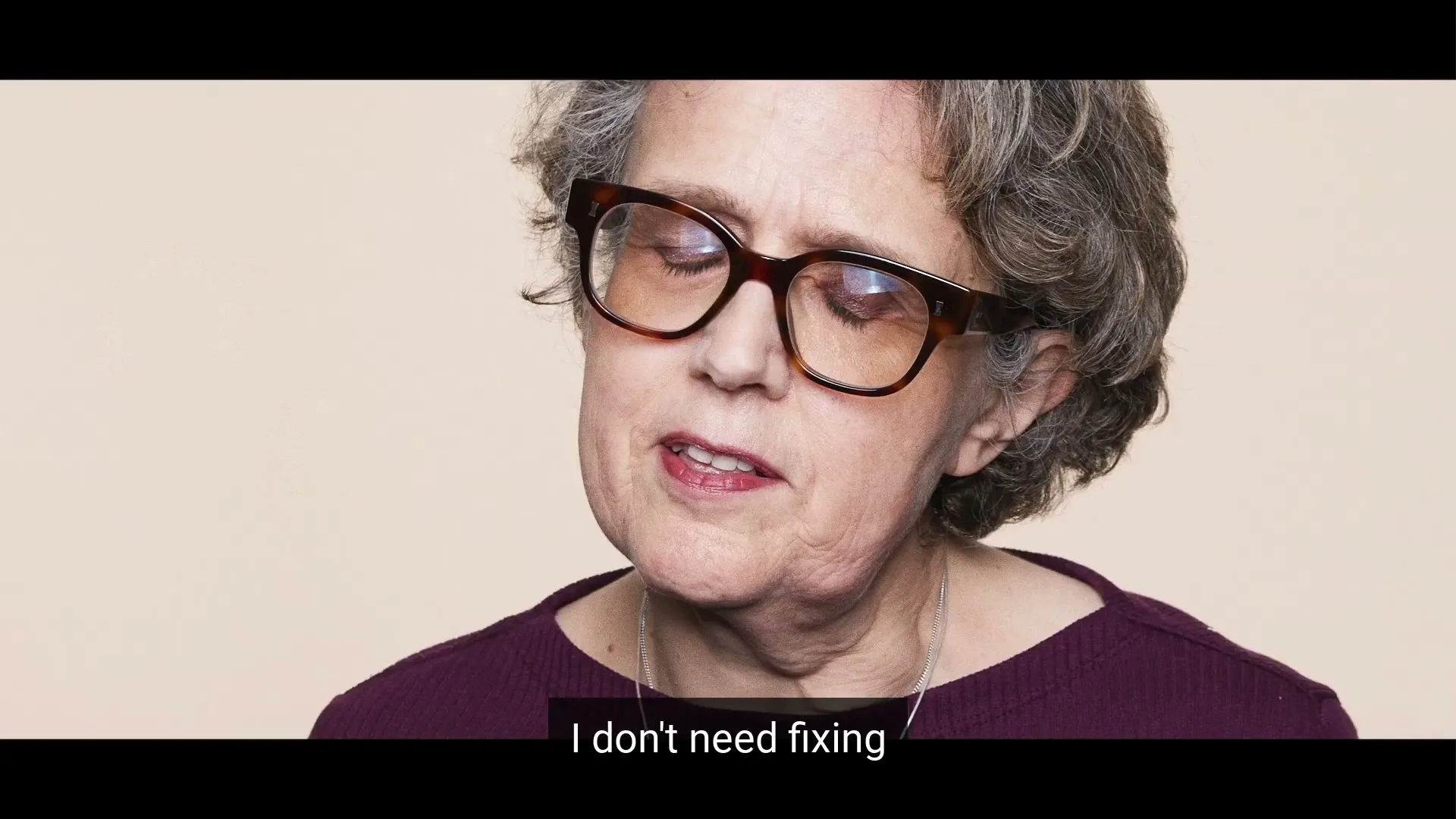 A woman with glasses and curly grey hair looks down and to the right, speaking with a slight smile against a plain background.