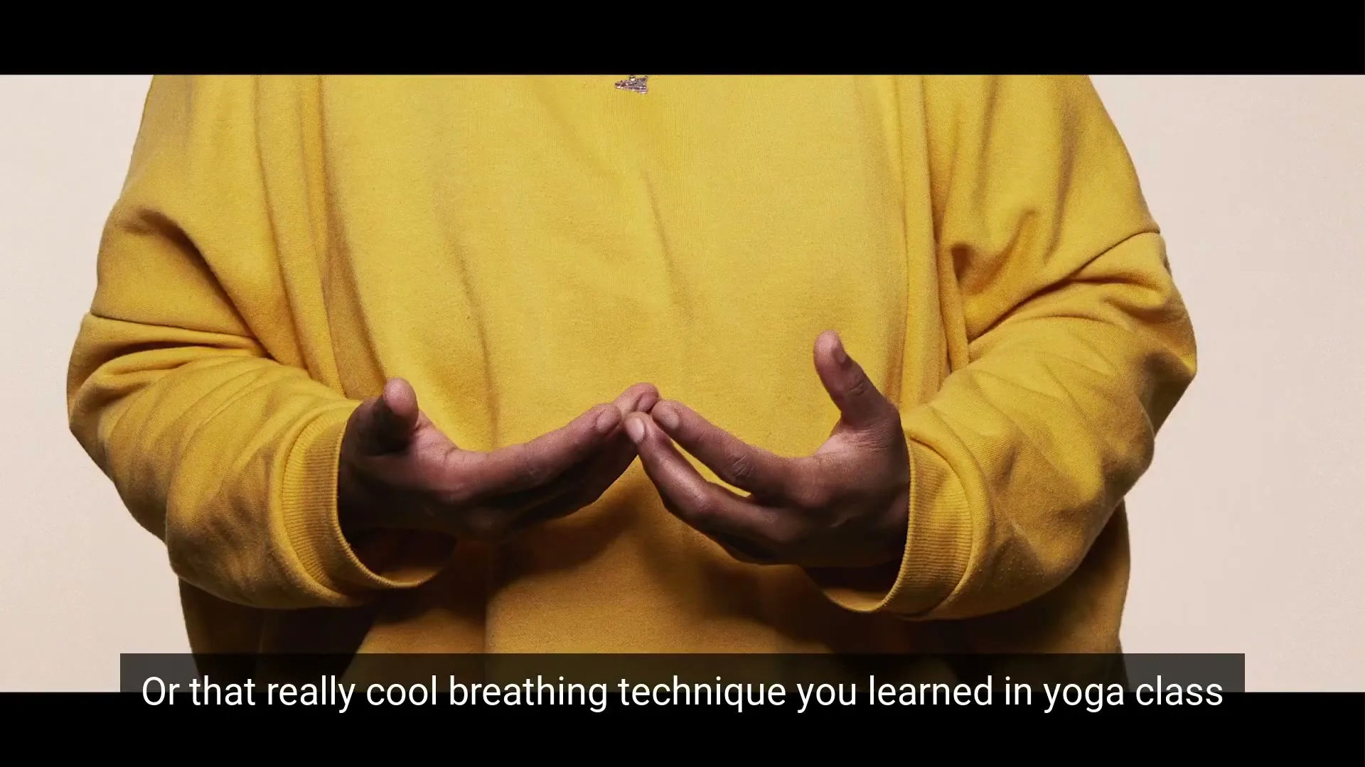 A person in a yellow sweatshirt demonstrates a breathing technique with their hands in a yoga class.