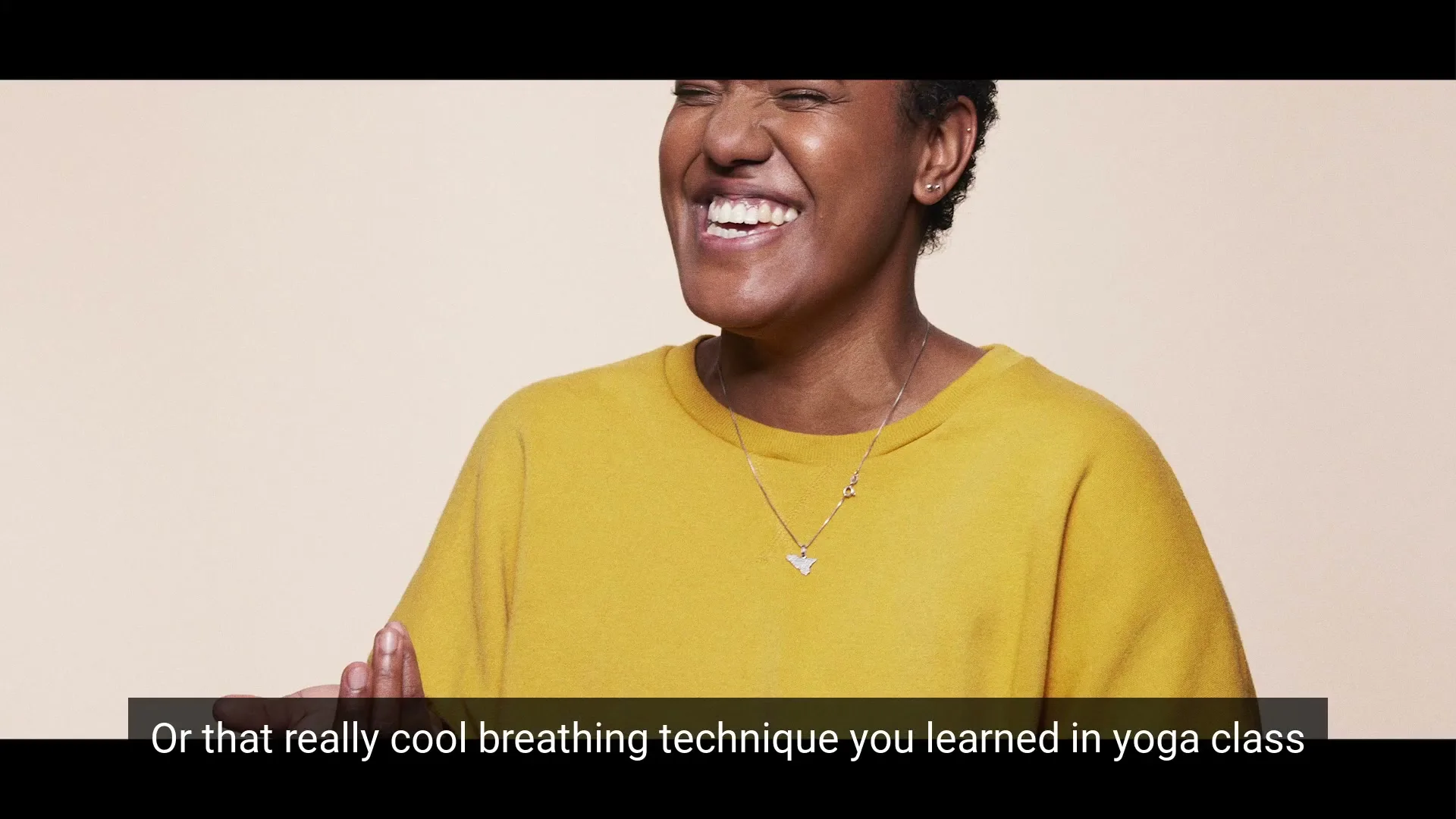 A Black woman in her 30s, wearing a yellow sweatshirt, laughs joyfully against a plain beige background.