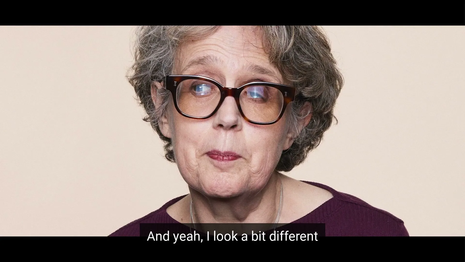A woman with grey curly hair and glasses looks to the side, wearing a purple top against a beige background.