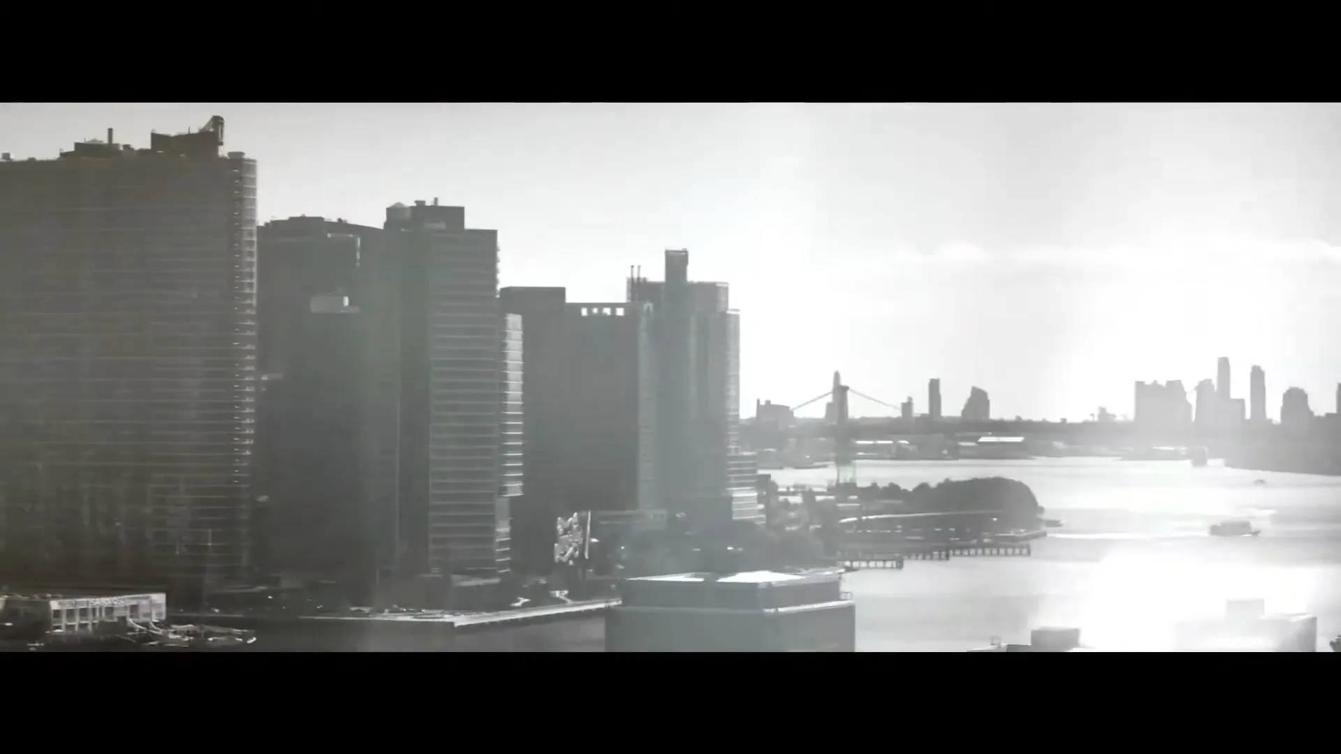 A black and white wide shot of the New York City skyline with tall buildings and a river, under a bright sky.