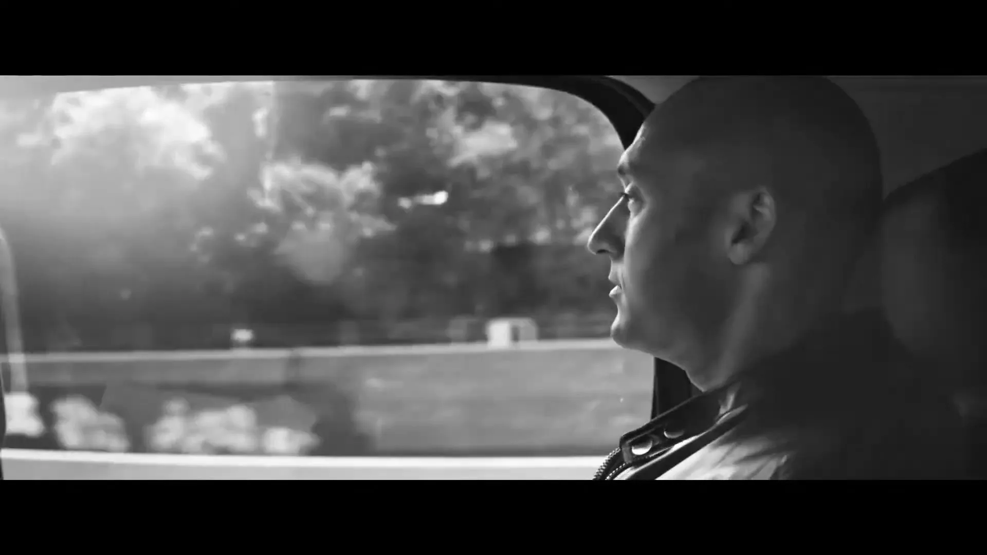 A bald man in his 30s, wearing a dark jacket, looks out the window of a moving car, with trees and buildings passing by in the background.