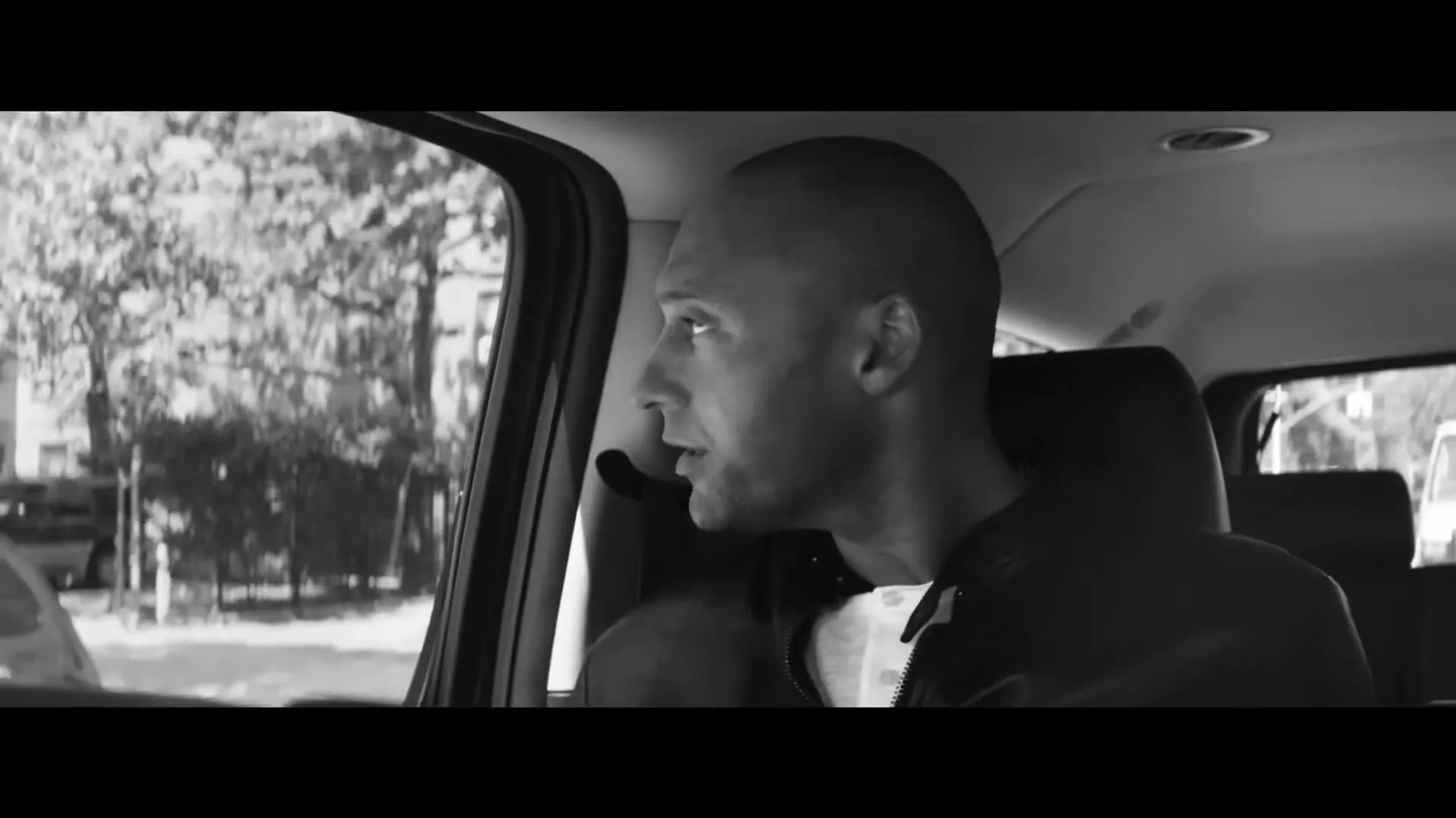 A bald man in his 30s, wearing a dark jacket, looks out the window of a moving car with a neutral expression.