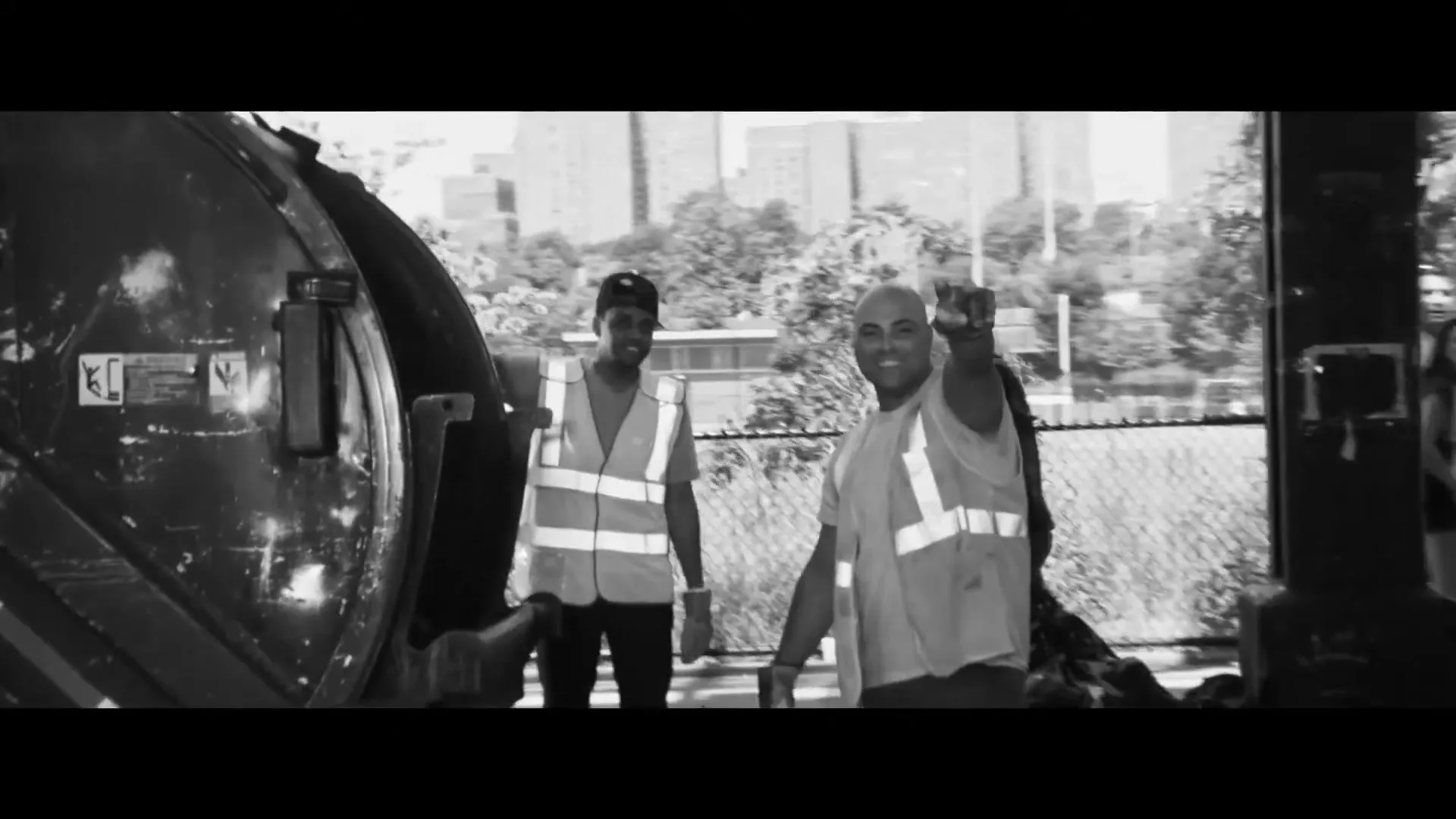 Two male sanitation workers in reflective vests stand next to a garbage truck, one pointing at the camera, on a sunny day in a city.