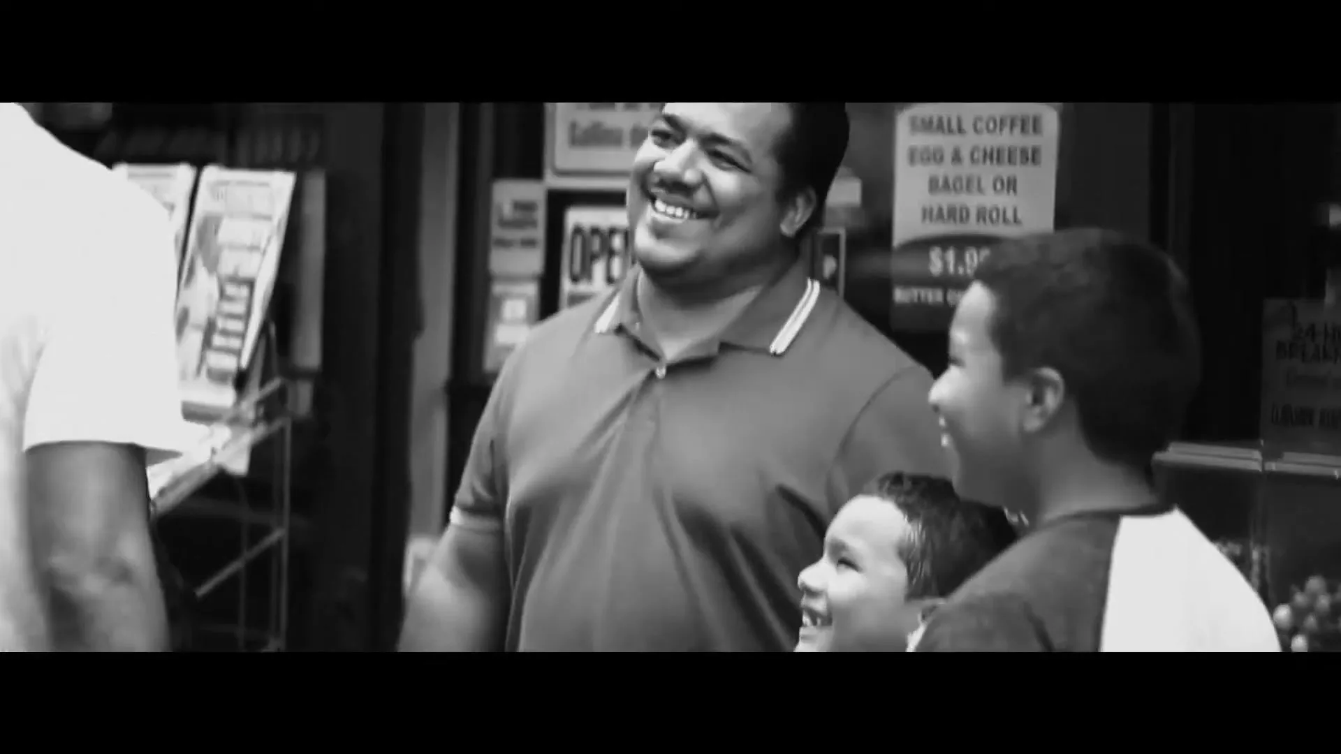A man in a polo shirt smiles with two young boys, one looking up at him, outside a storefront in black and white.