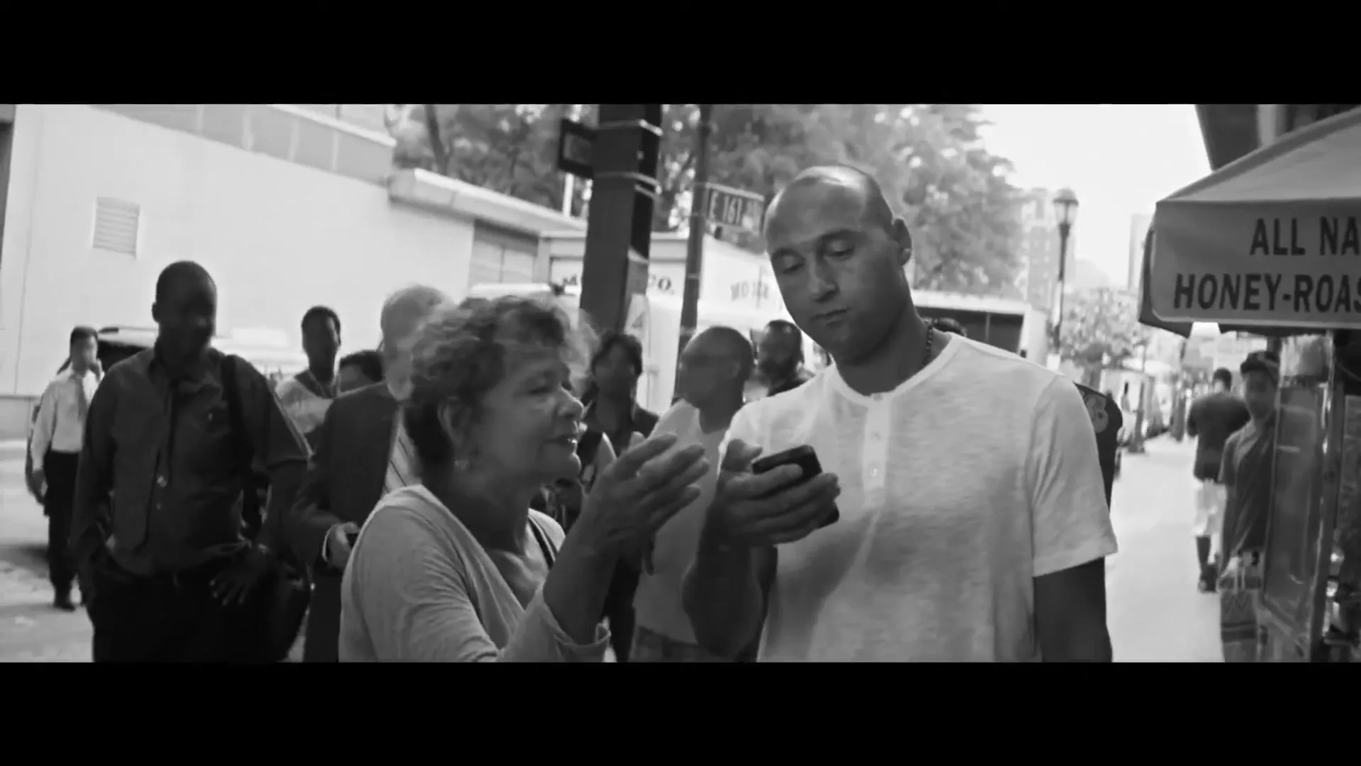 Derek Jeter, a bald man in a white shirt, shows his phone to an older woman with curly hair on a busy New York City street.