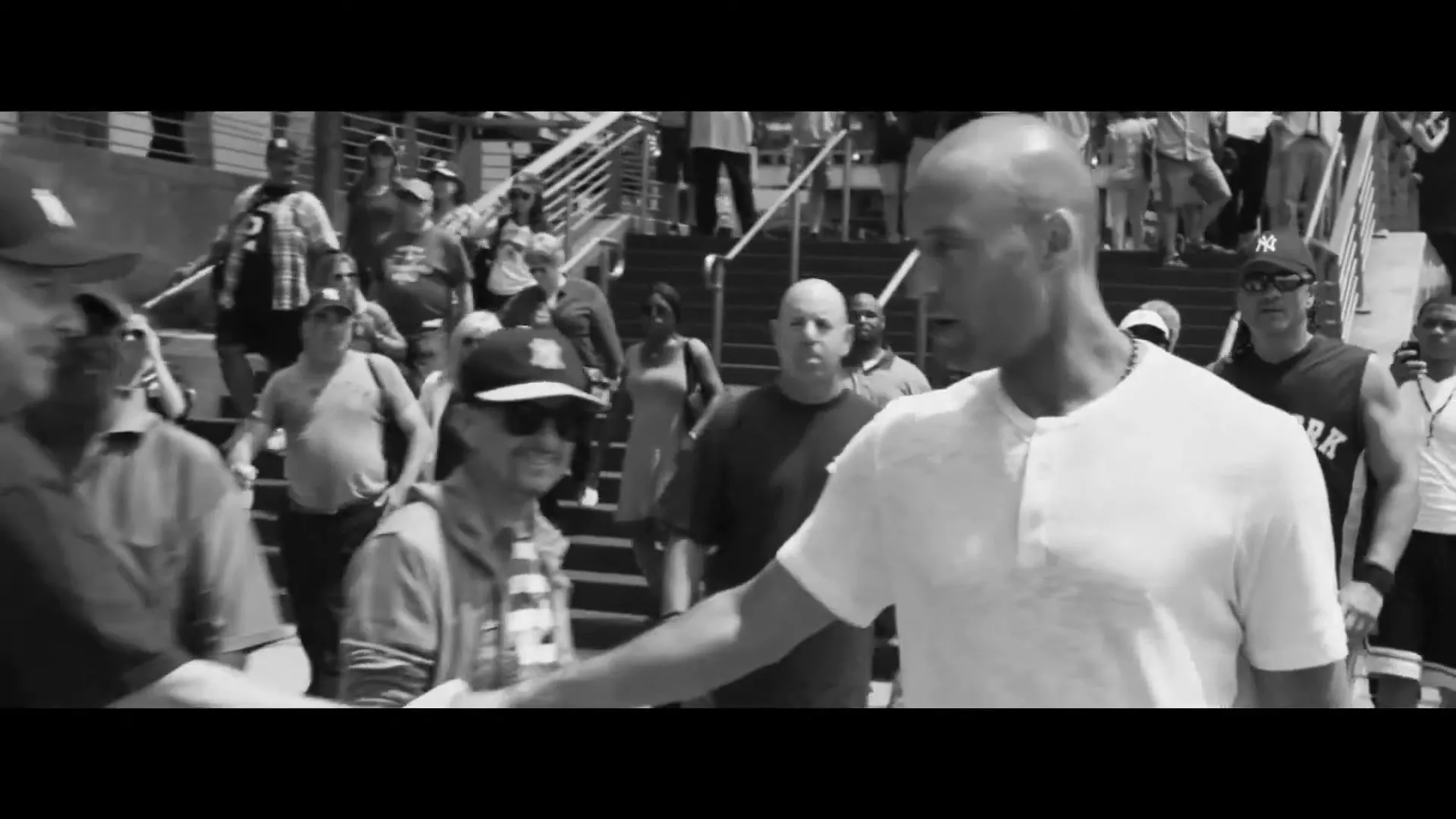 A bald man in a white shirt shakes hands with a fan while walking through a crowd of people on a sunny day outside a stadium.