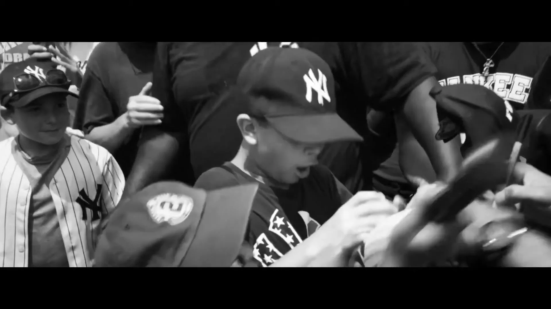A young boy in a Yankees cap and t-shirt is surrounded by a crowd, excitedly receiving an autograph on a baseball.