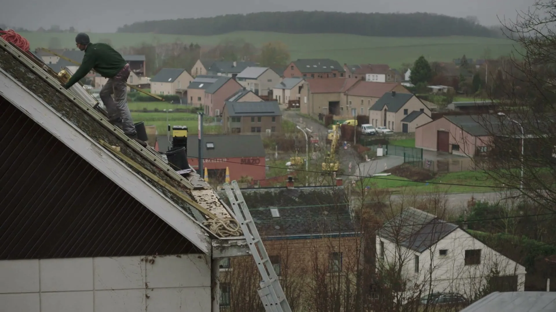 A male roofer in a green jacket and grey pants works on a steep roof, with a village and green hills in the background on a cloudy day.
