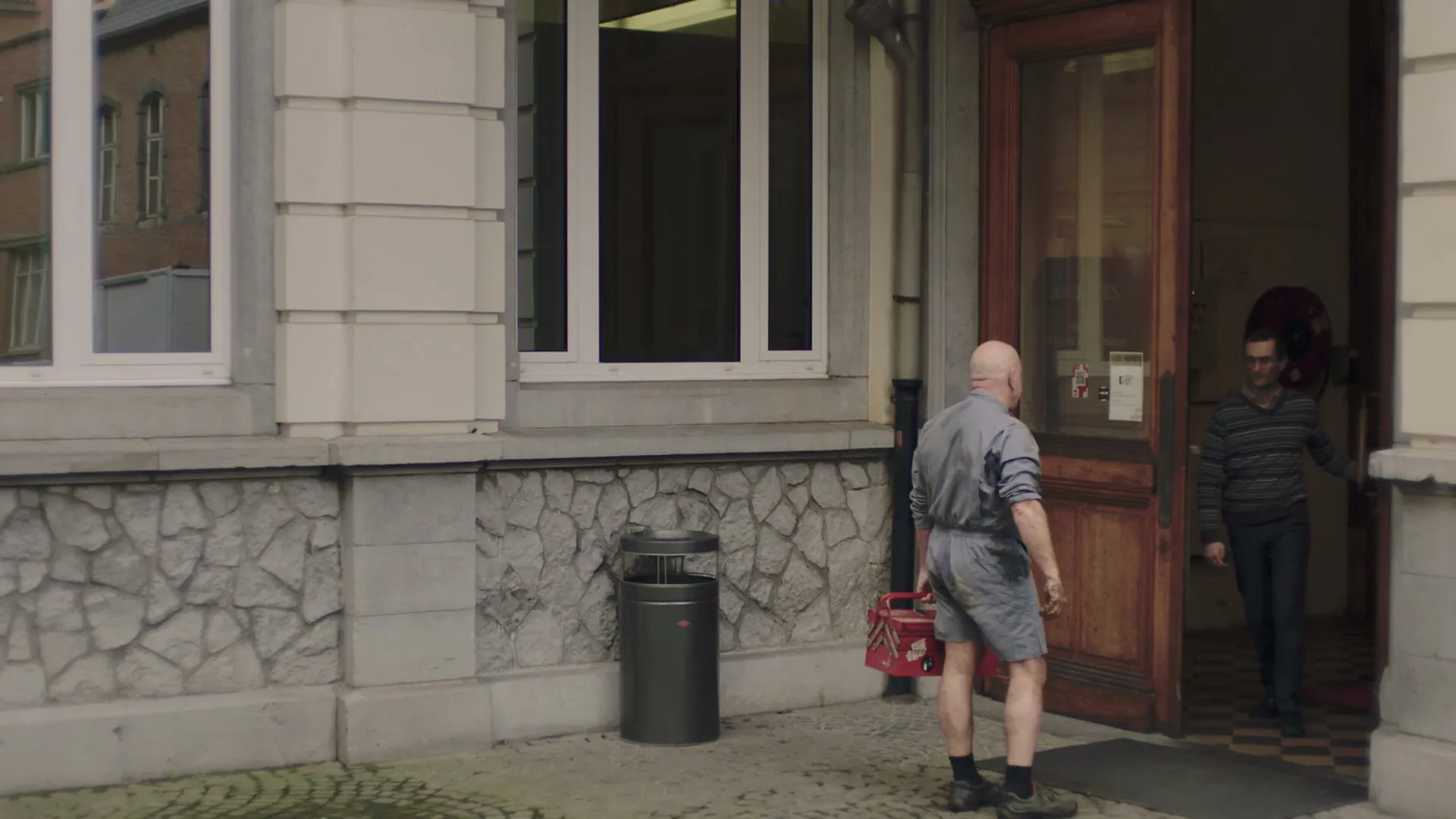 A bald man in dirty work clothes holds a red toolbox and stands outside a building as another man exits through a wooden door.