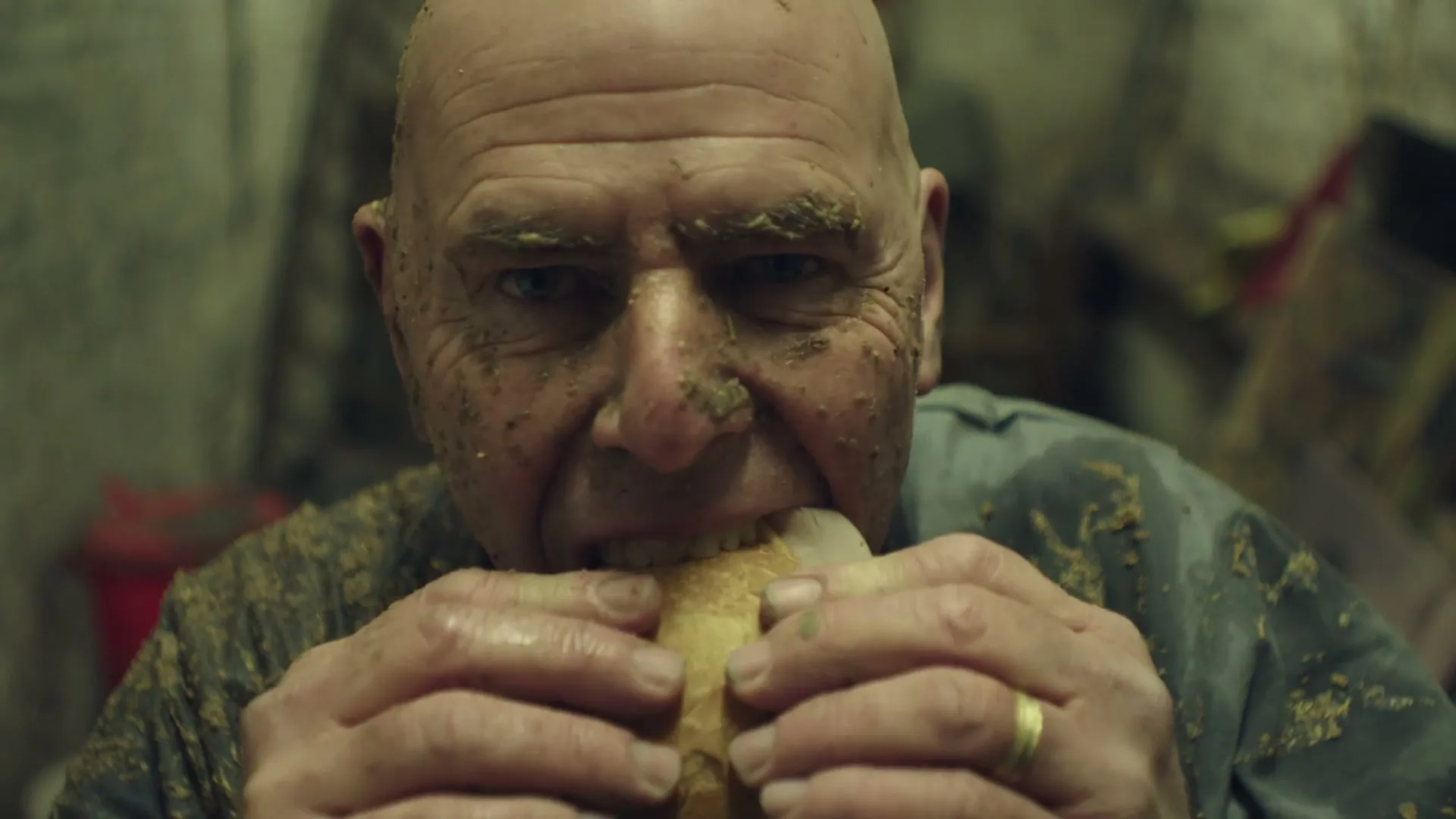 A bald man, covered in dirt and grime, aggressively eats a sandwich in a dimly lit, messy interior.