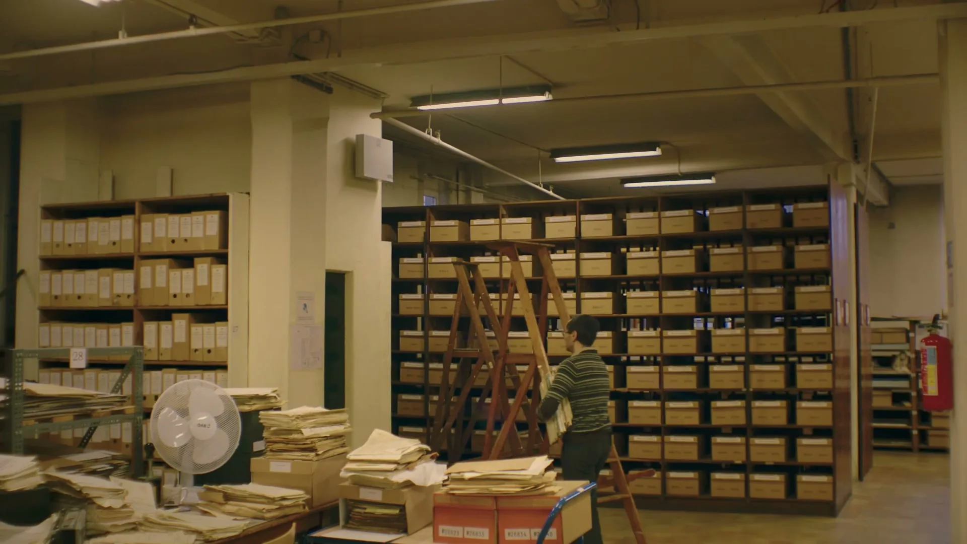 A man in a striped shirt and dark pants carries a stack of papers while standing on a wooden ladder in a cluttered archive room.