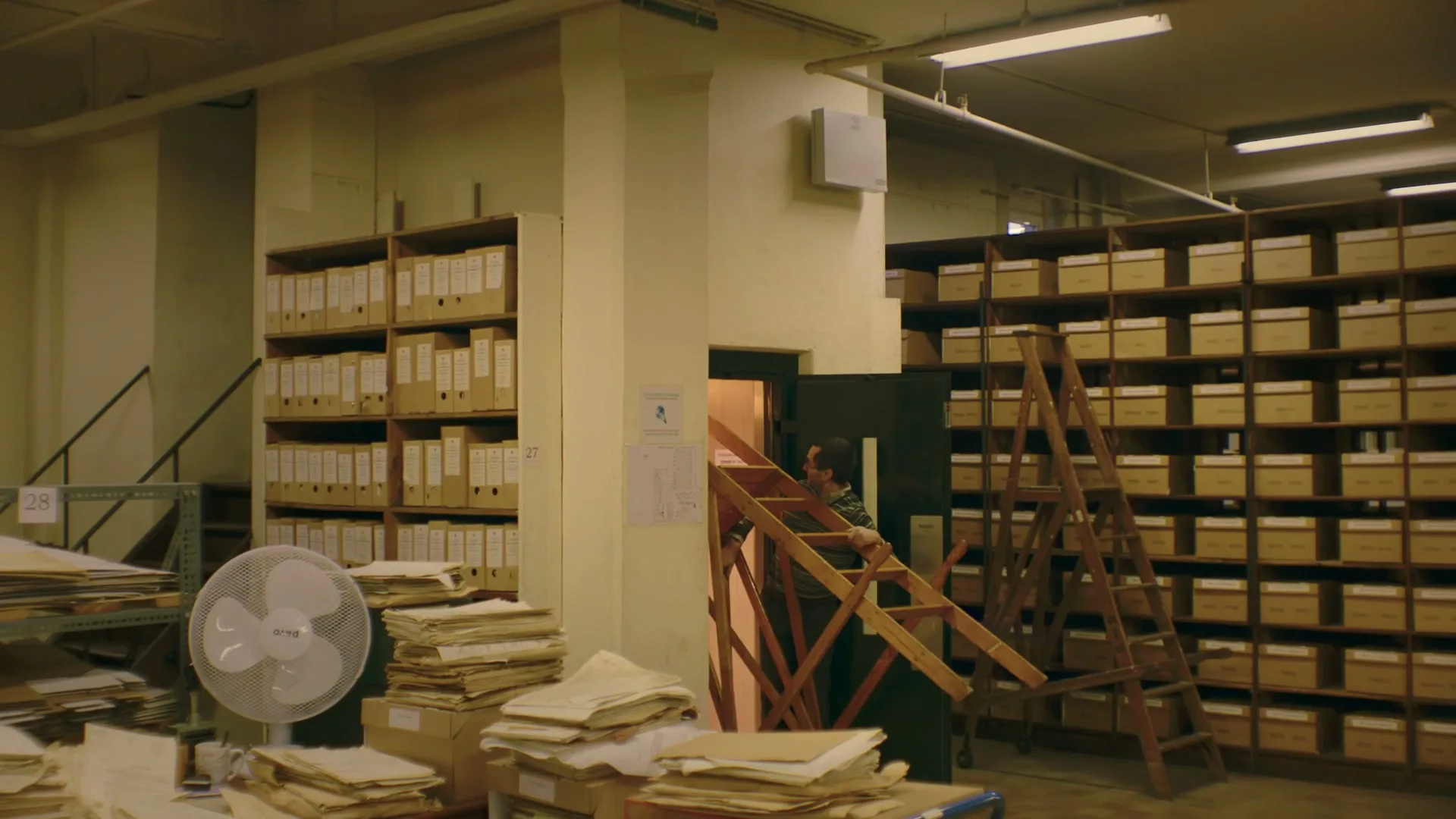 A man in a green shirt and dark pants is seen through a doorway, moving a wooden ladder in a cluttered archive room filled with shelves of files and boxes.
