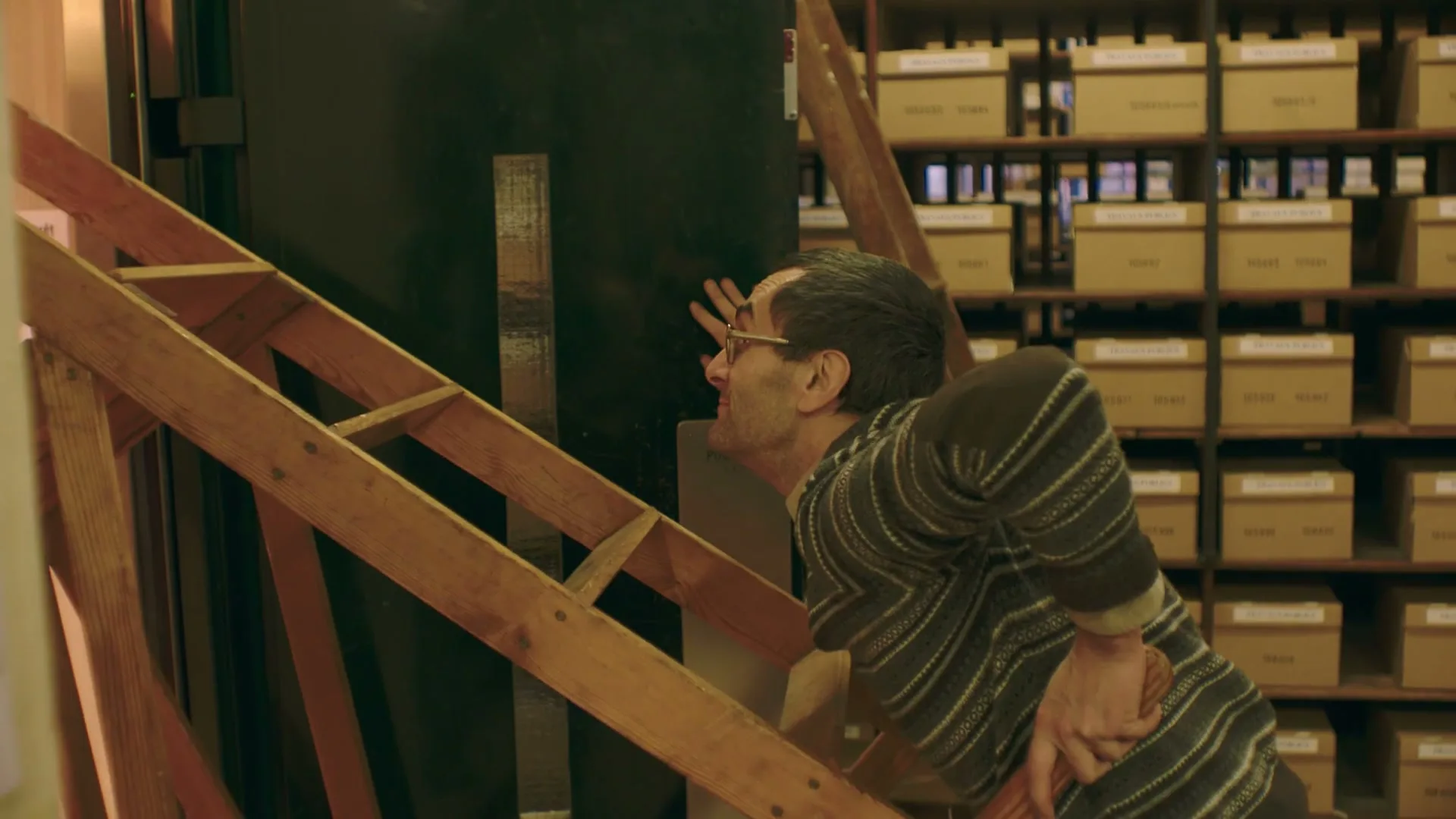 A man in glasses and a striped sweater is seen from the side, leaning against a dark surface with a wooden ladder in the foreground, in a room with shelves of boxes.