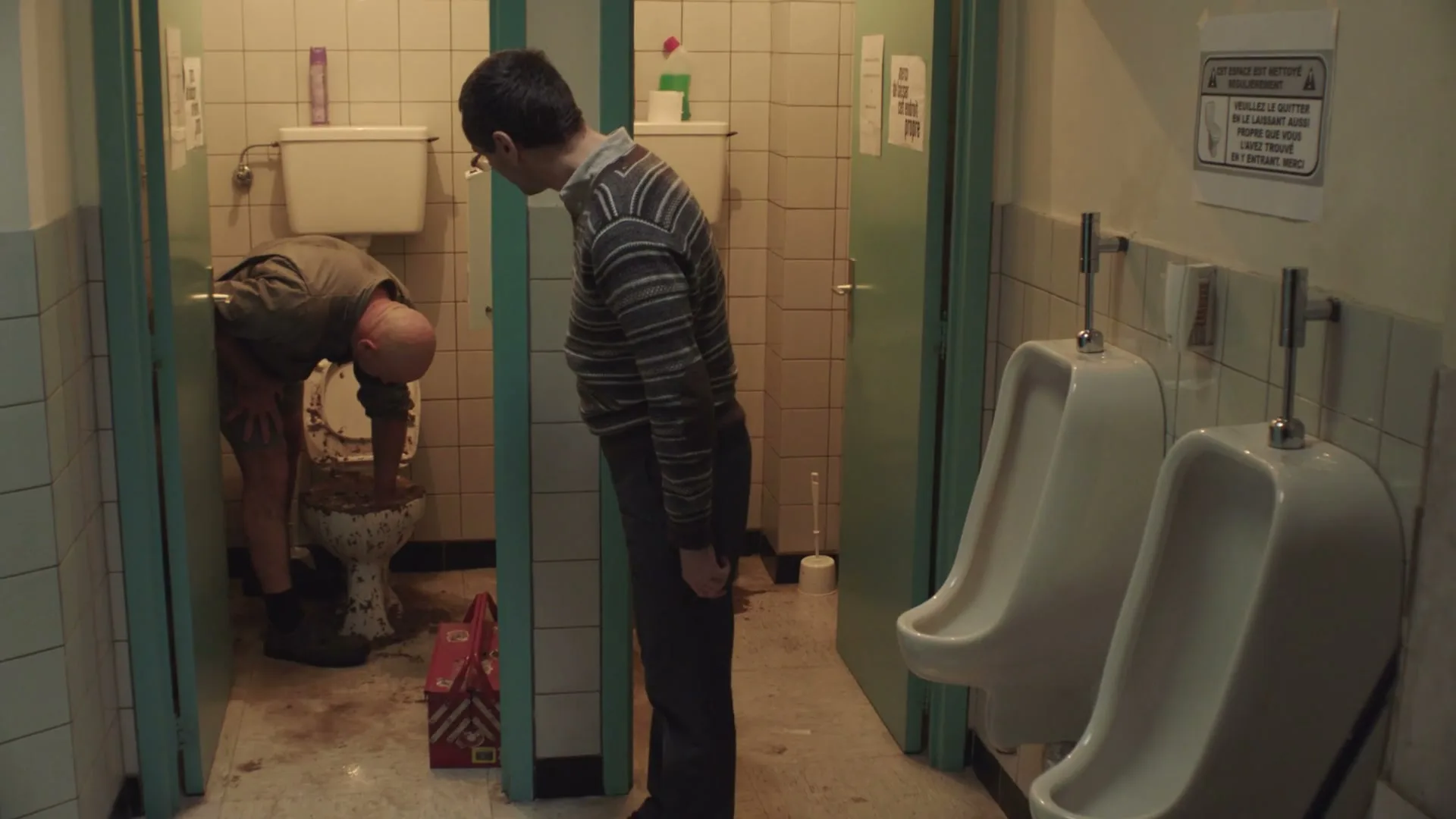 A bald man in a dirty uniform cleans a disgustingly overflowing toilet while another man in glasses watches him in a public restroom.