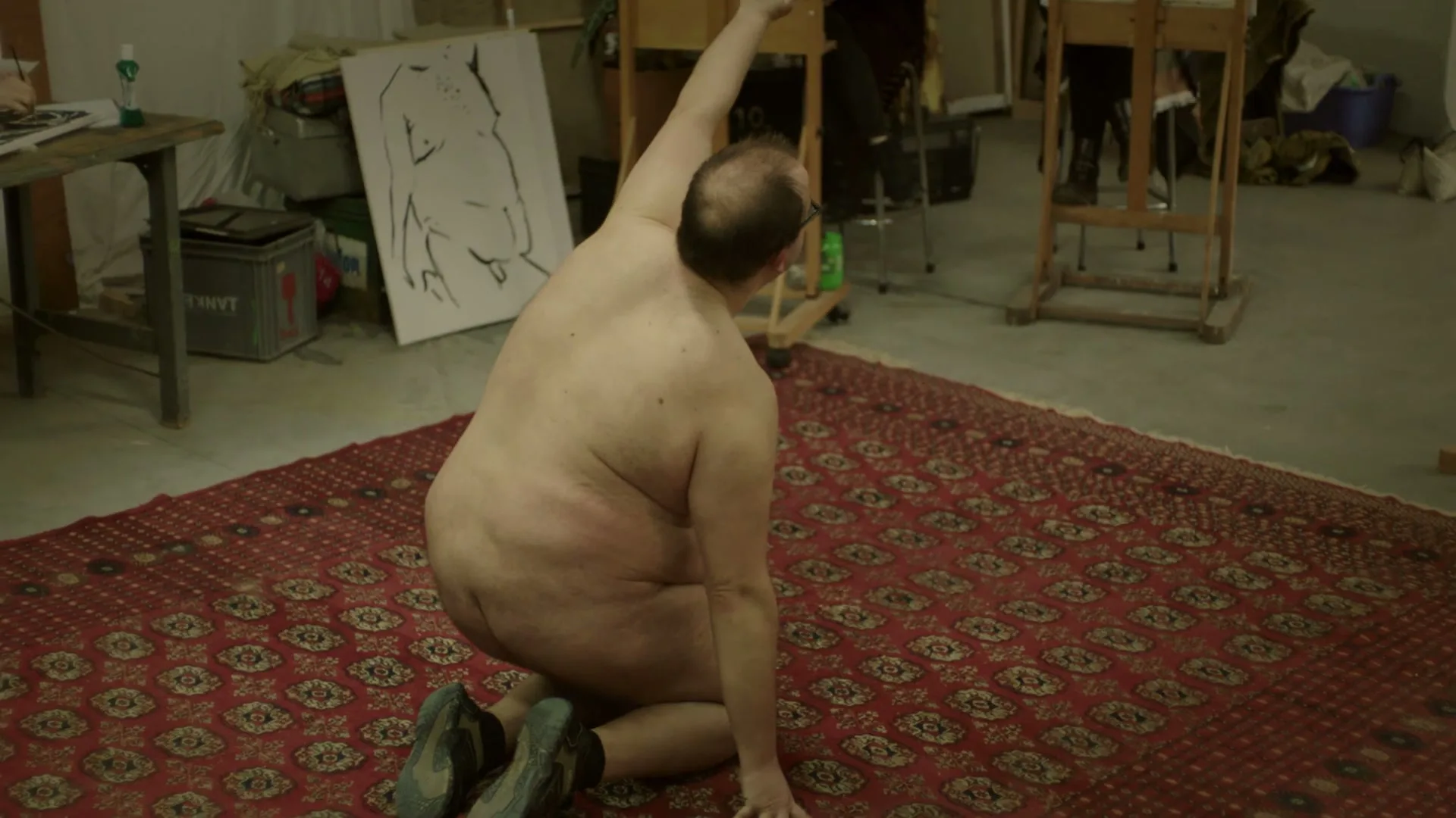 A bald, nude man with glasses kneels on a red patterned rug, stretching his arm upwards in an art studio.