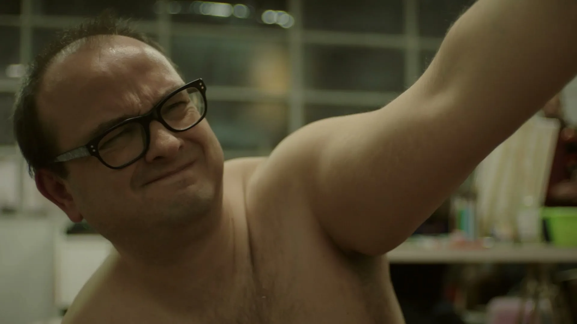 A man with glasses and a bare chest grimaces while raising his arm, possibly applying deodorant, in an interior setting.