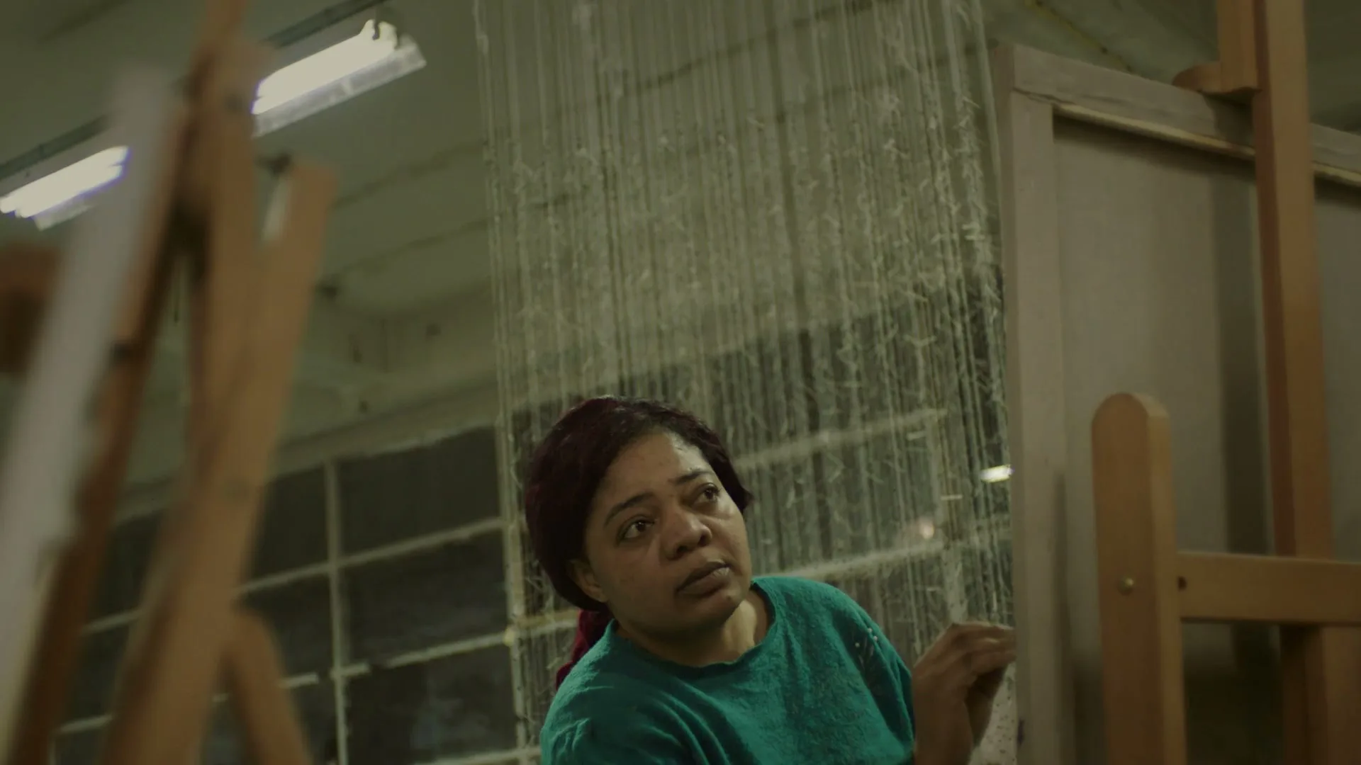 A woman with dark red hair and a teal shirt looks up while working on a loom in a dimly lit studio.