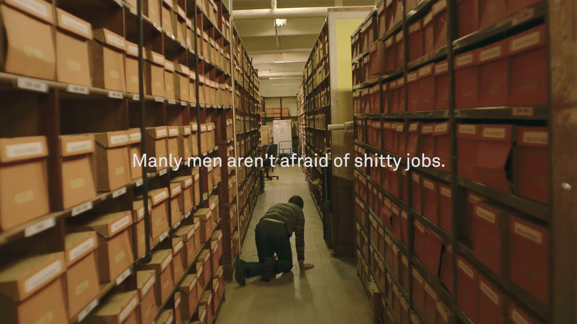 A man in a striped sweater and dark pants crawls on his hands and knees down a narrow aisle between tall shelves filled with cardboard boxes in an archive.