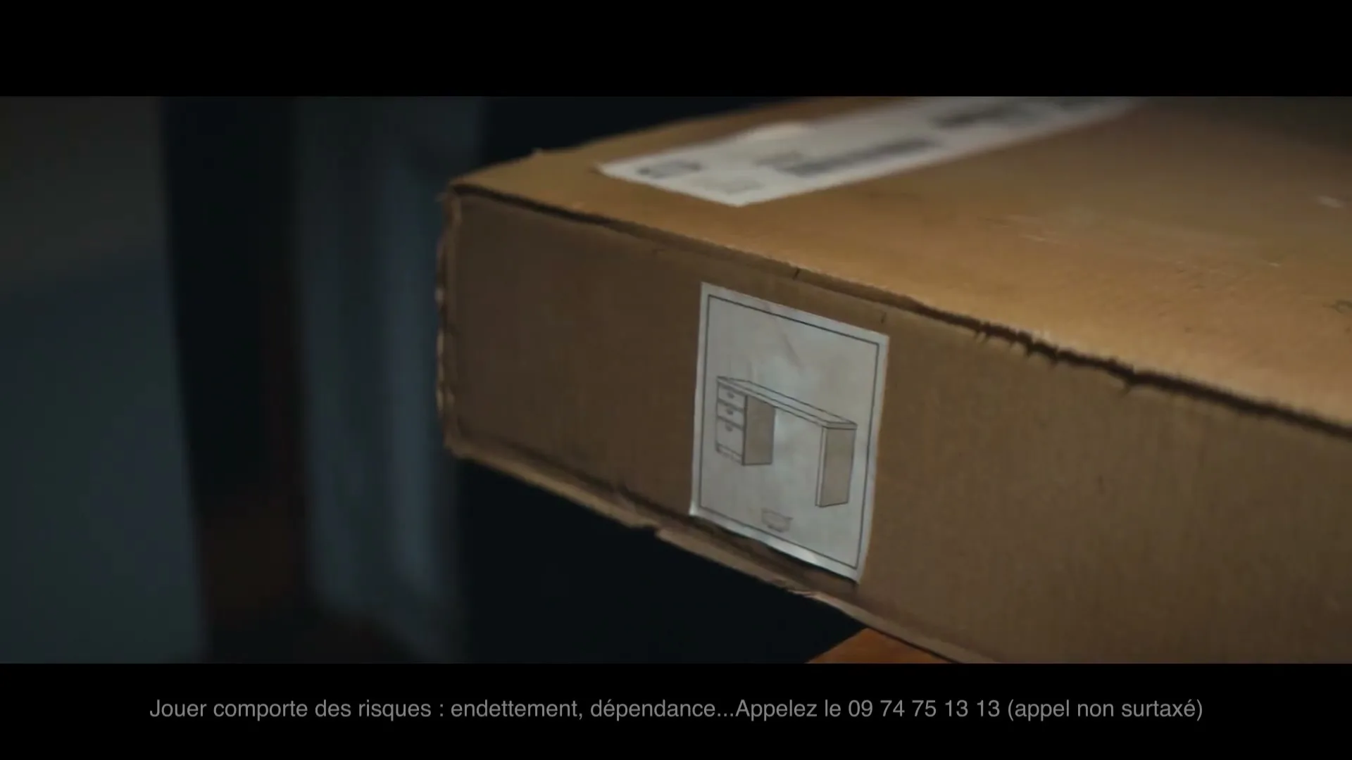 A close-up shot of a cardboard box with a label showing a diagram of a desk, sitting on a surface in an interior setting.