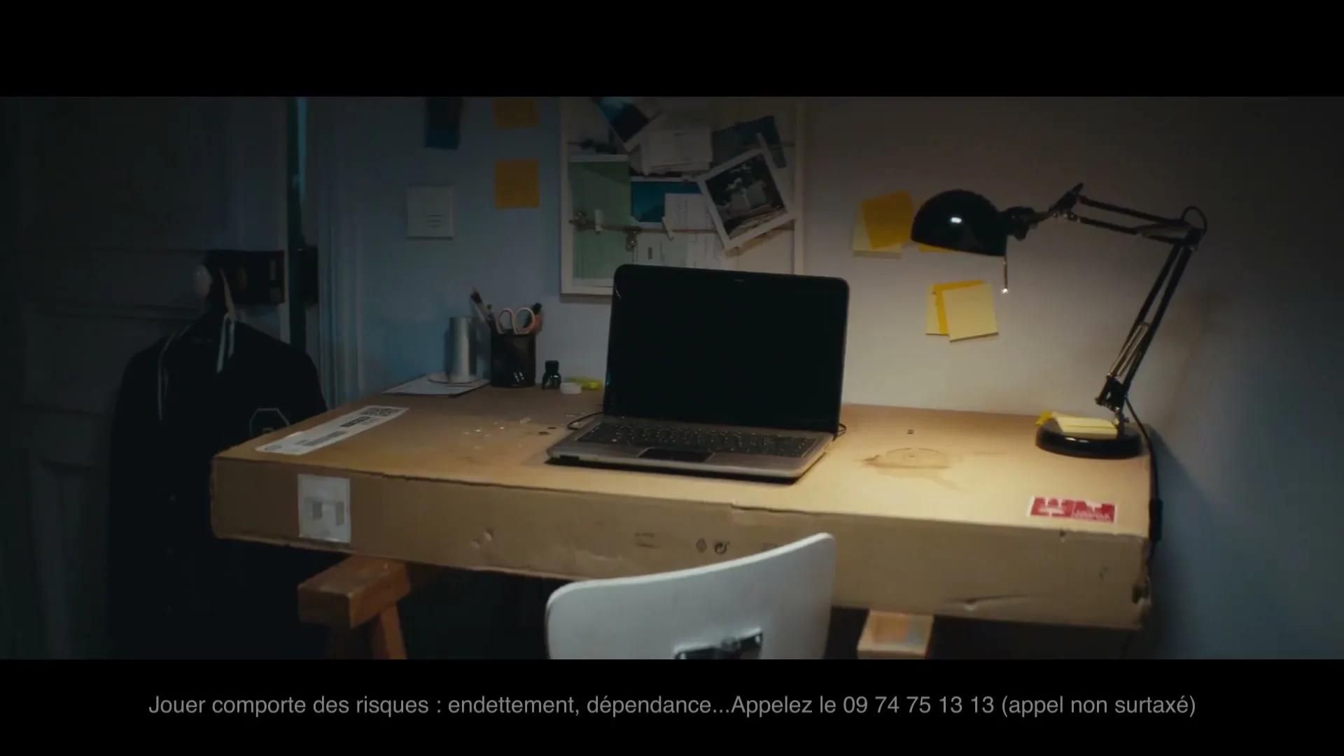 A laptop sits on a makeshift cardboard desk in a dimly lit room, with a desk lamp illuminating the workspace.