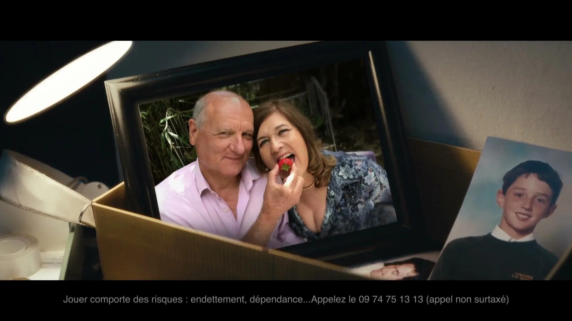 A framed photo of a smiling older couple, with the woman eating a strawberry, is placed in a cardboard box with other photos.