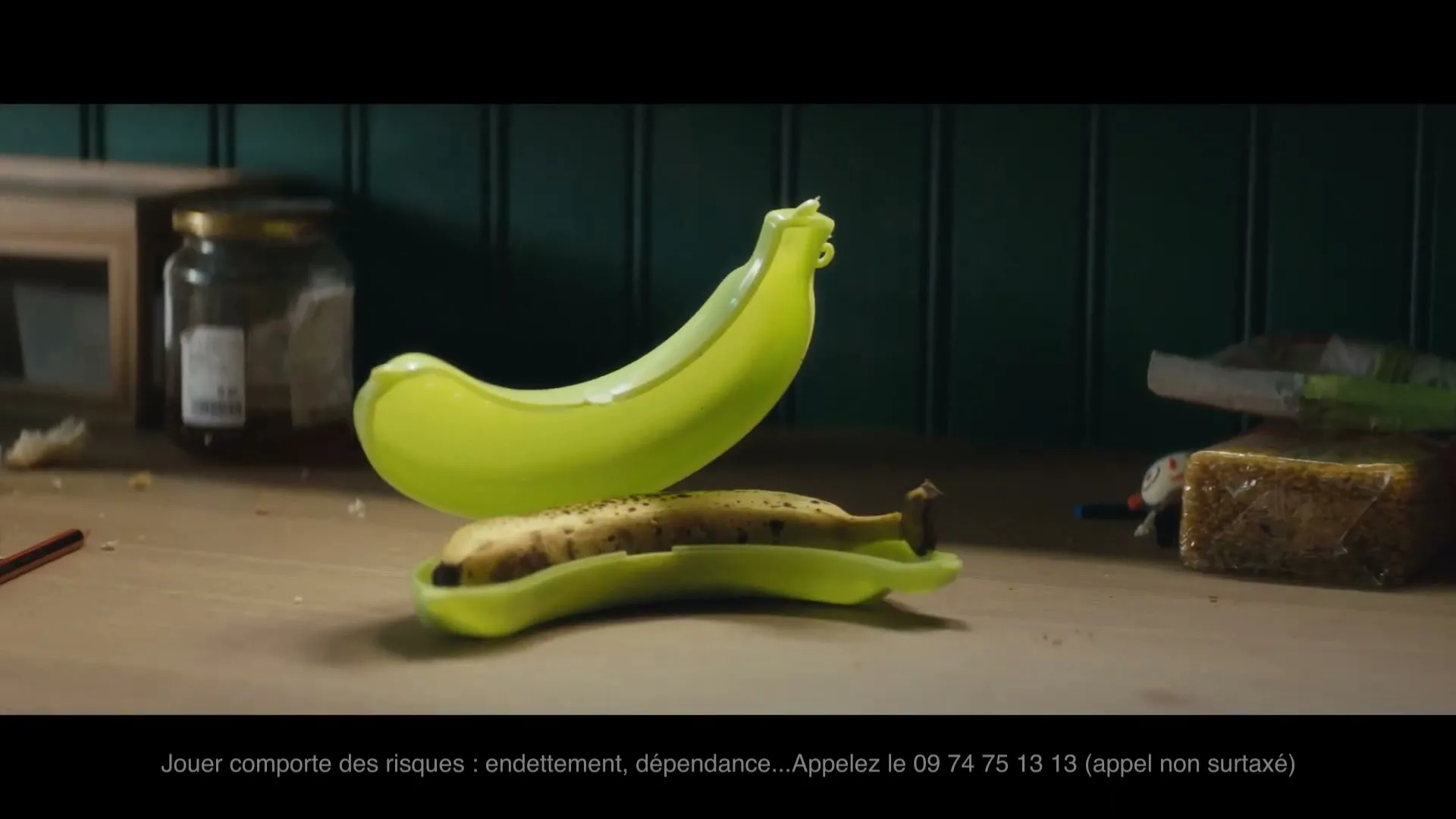 A banana in a plastic banana case sits on a messy wooden table, with a jar and other items in the background.