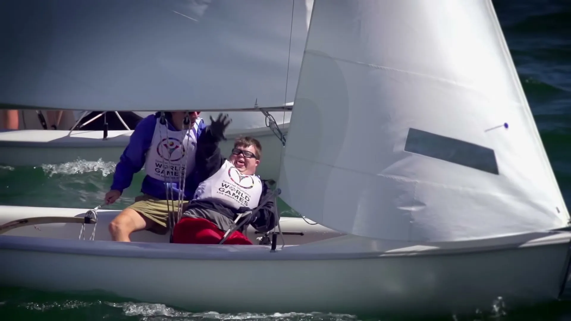 Two men, one with a disability, are sailing a small white sailboat on a sunny day, both wearing "World Games" vests.