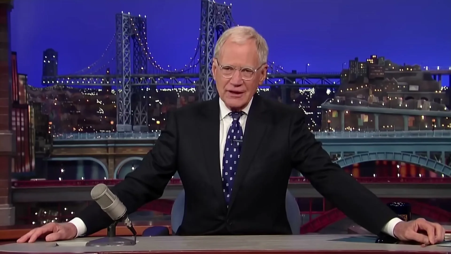 David Letterman, a white man in his 60s, wearing a suit and tie, sits at a desk on a late-night talk show set, smiling.