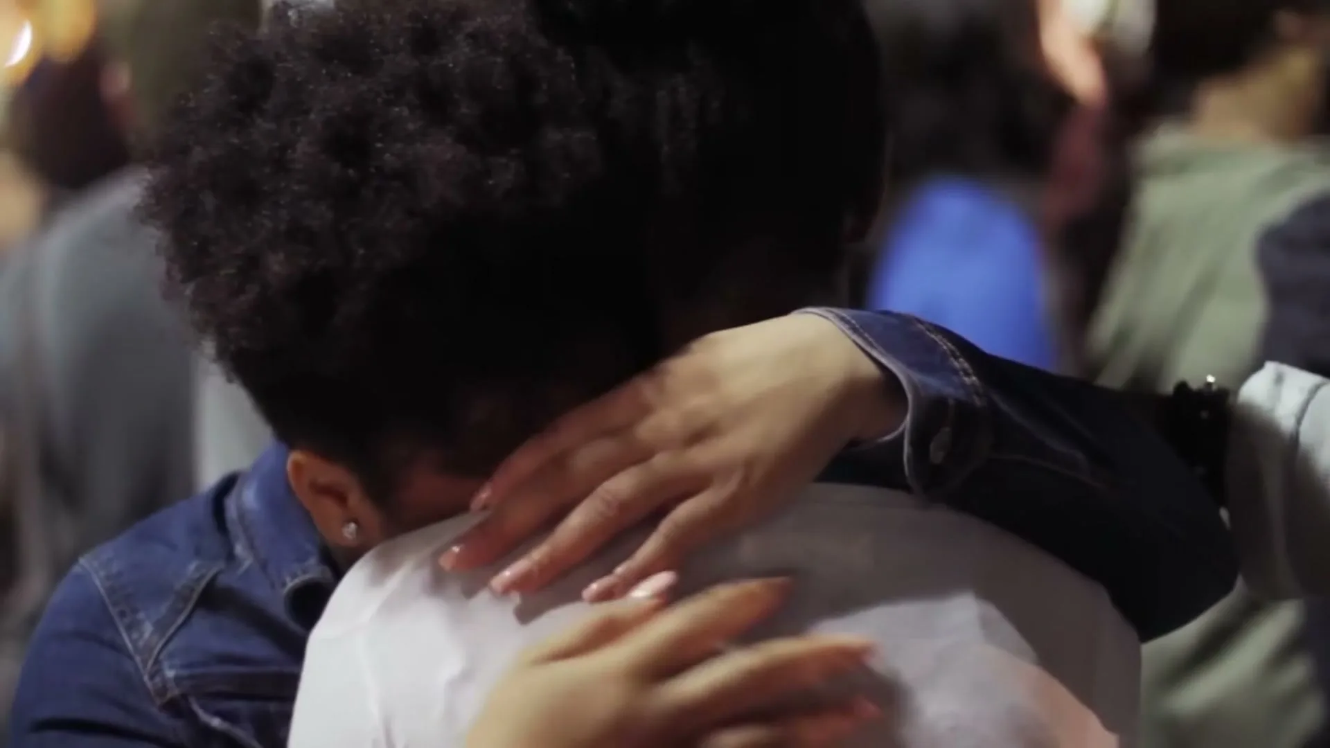 Two people with dark curly hair embrace tightly, one wearing a denim jacket and the other a white shirt, in a crowd of people.